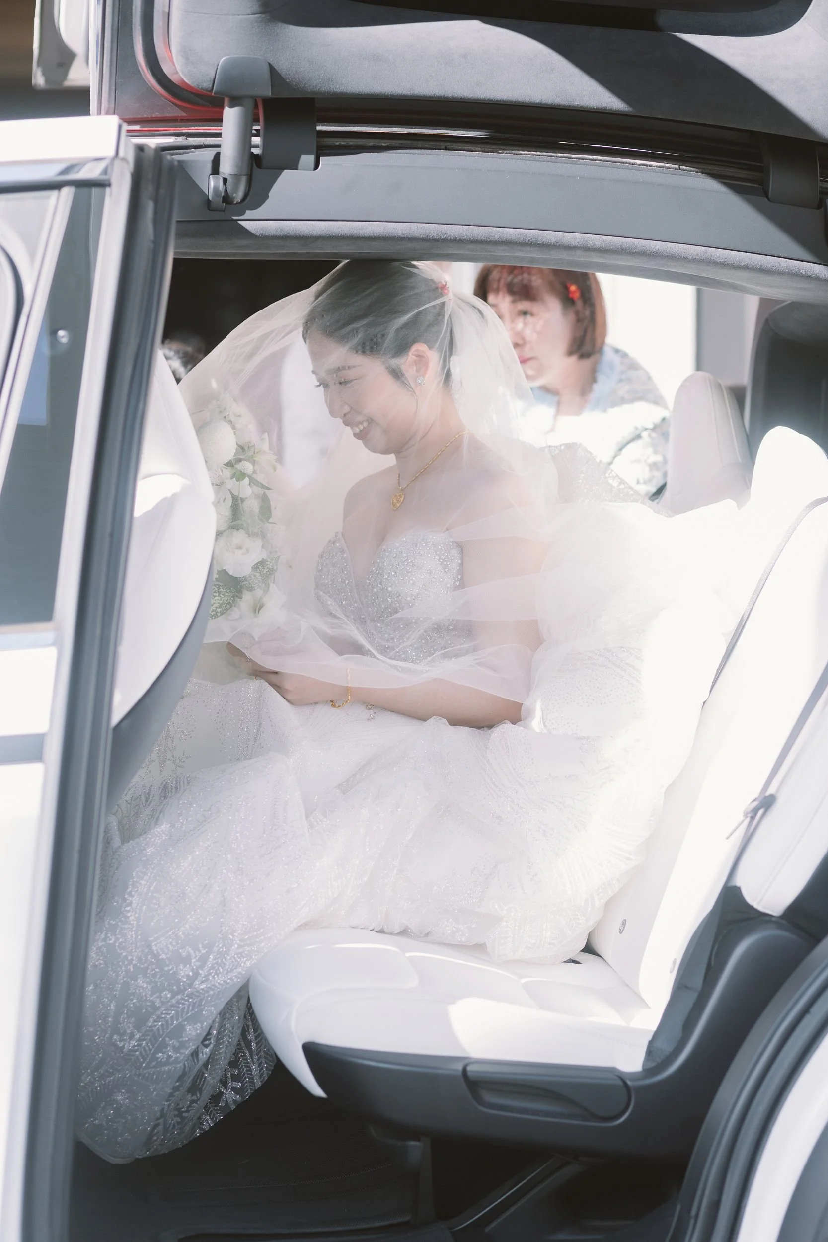 A bride in a wedding gown with a veil sitting in the back seat of a car, smiling with her eyes closed, holding flowers. A person behind her is looking over her shoulder. The photo is taken through the open door of the car.