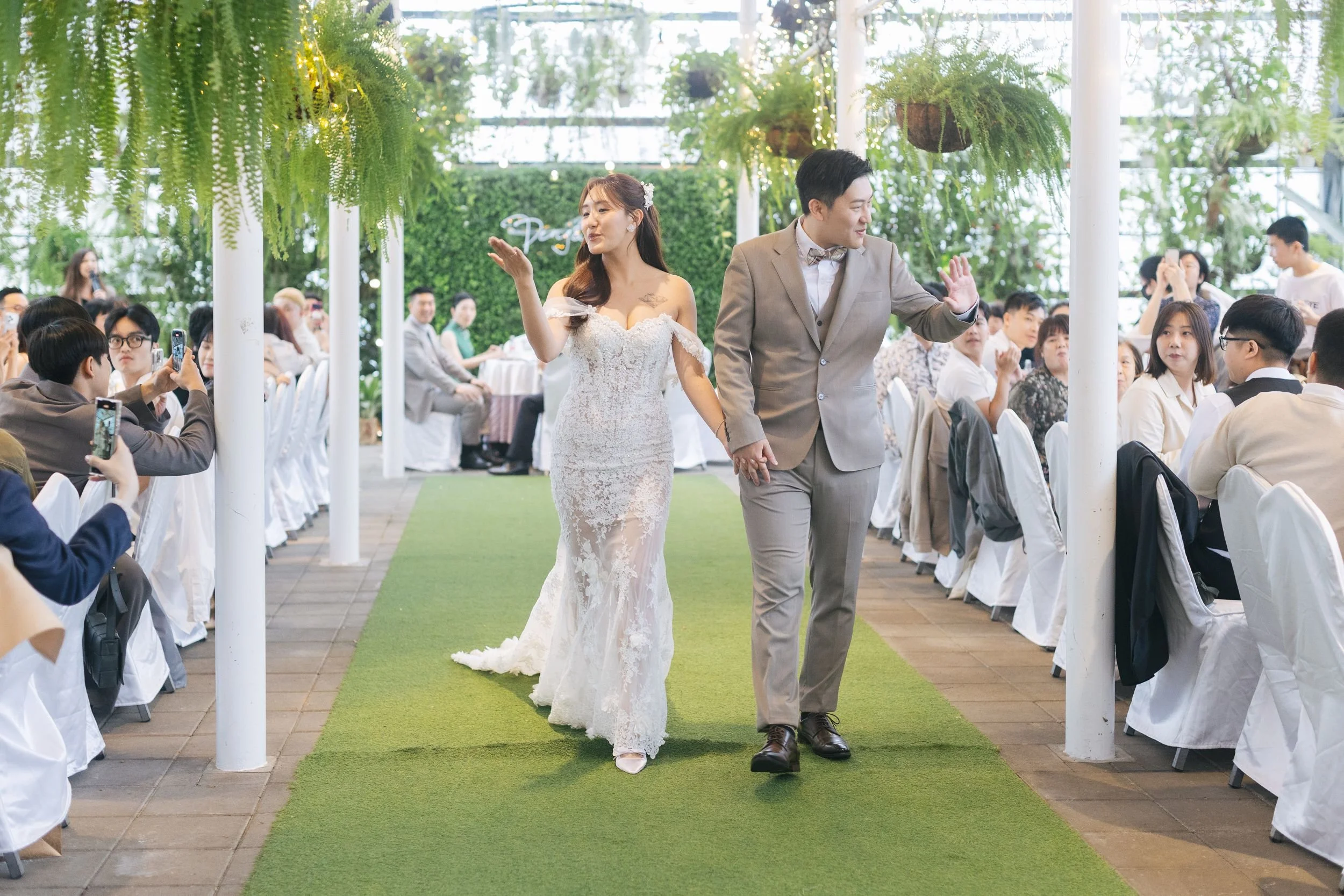 A bride and groom holding hands and walking down the aisle during a wedding ceremony in a decorated indoor venue with hanging plants and a green carpet.