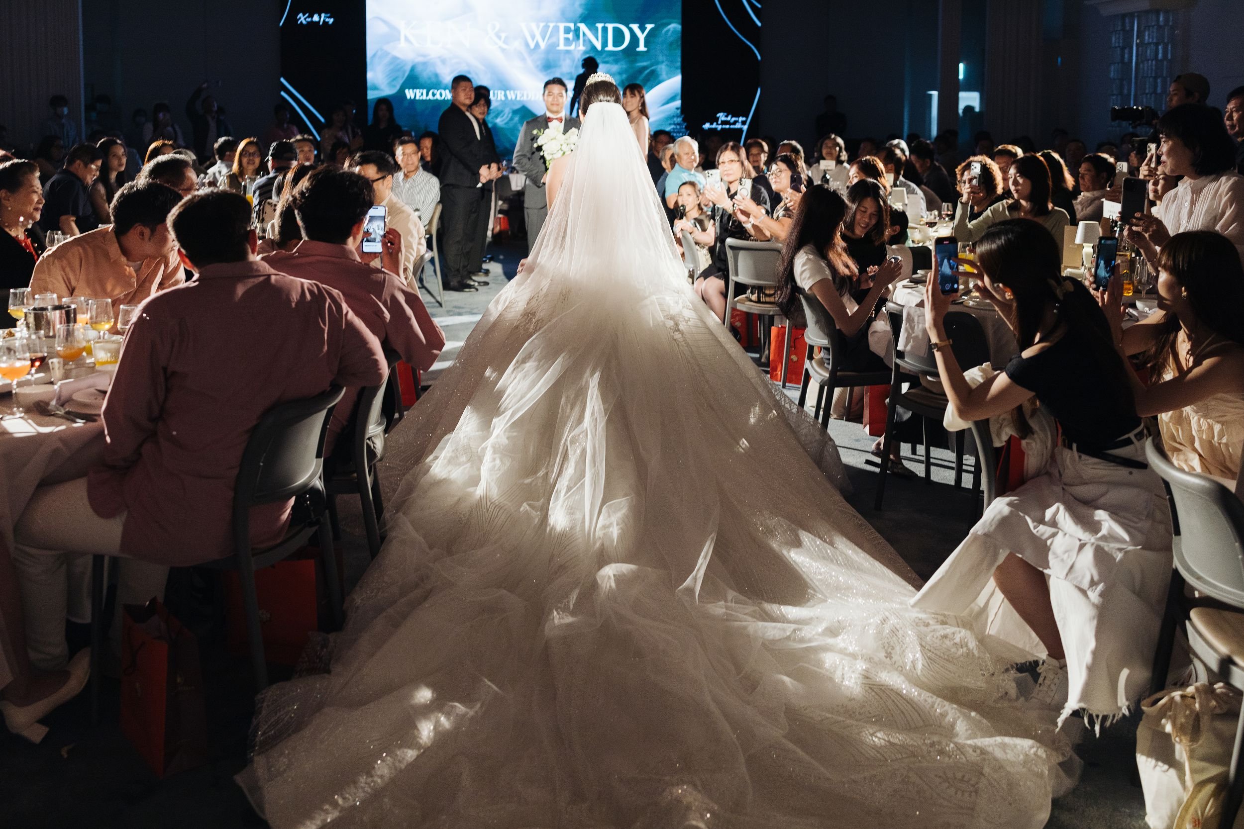 A bride in a wedding gown and veil walking down an aisle at her wedding reception with guests seated on both sides, many taking photos with their phones. A large digital screen in the background displays wedding-related text.