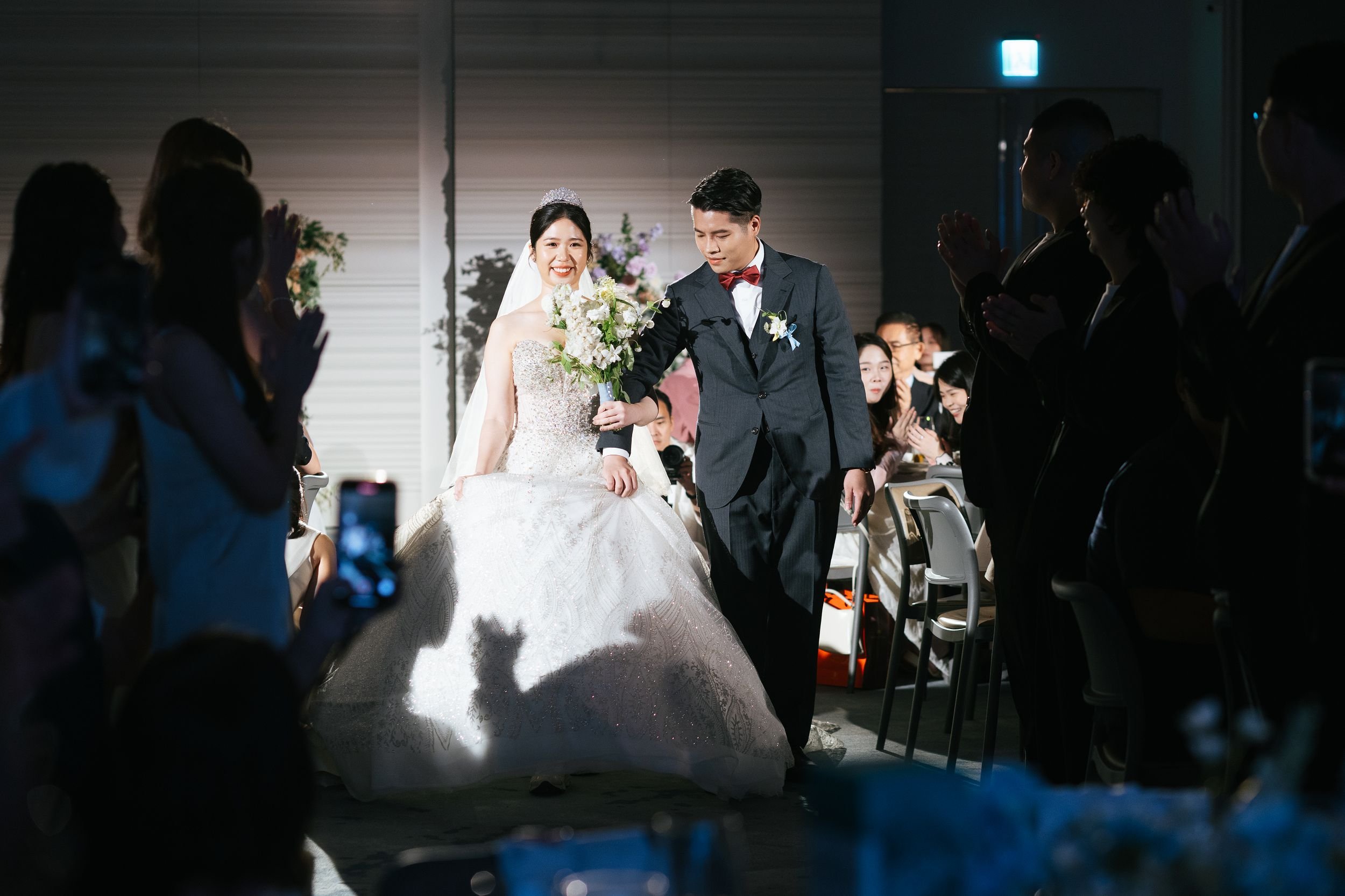 A bride and groom are walking down the aisle at their wedding, smiling. The bride holds a bouquet of flowers and wears a tiara, while the groom is dressed in a suit with a red bow tie. Guests are standing and clapping on either side in a dimly lit ro