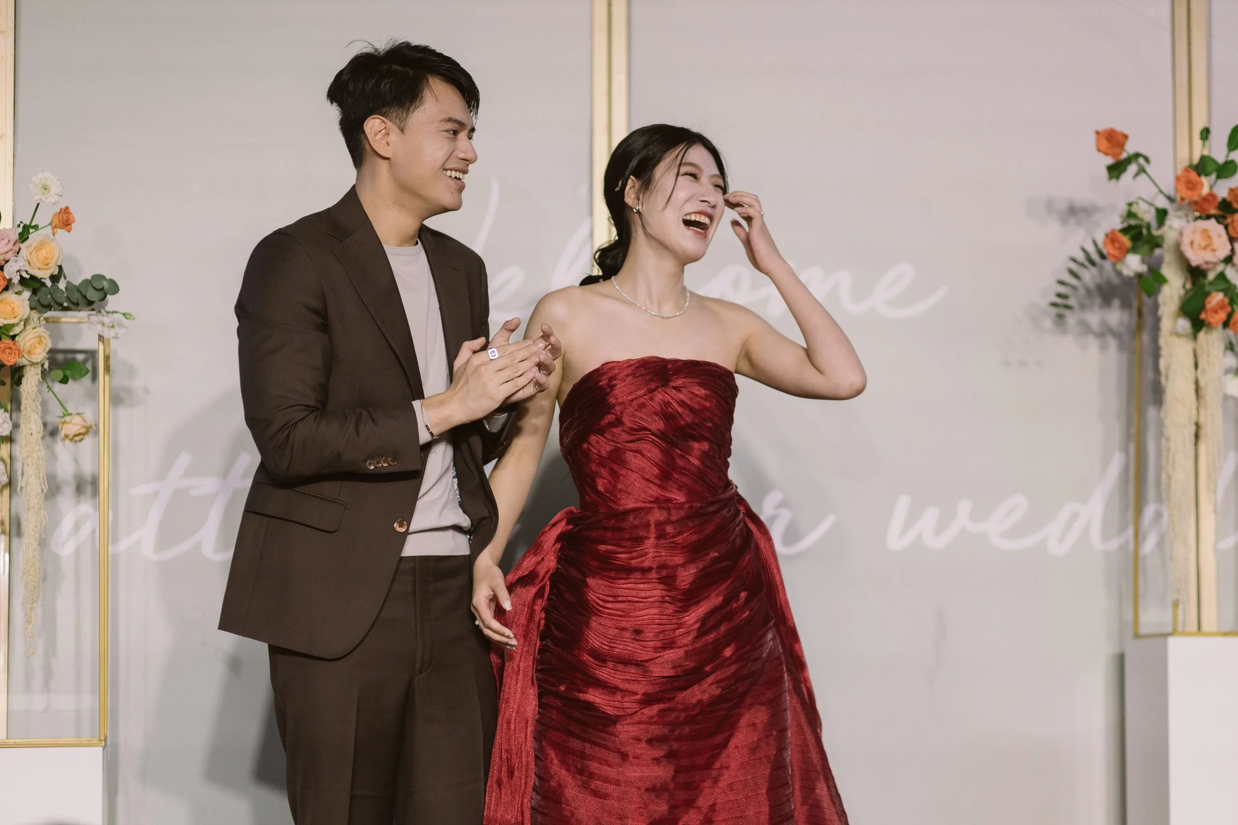 A young couple at a celebration, with the woman in a strapless red dress and the man in a brown blazer. They are smiling and laughing together, holding hands in front of a decorated backdrop with flowers.