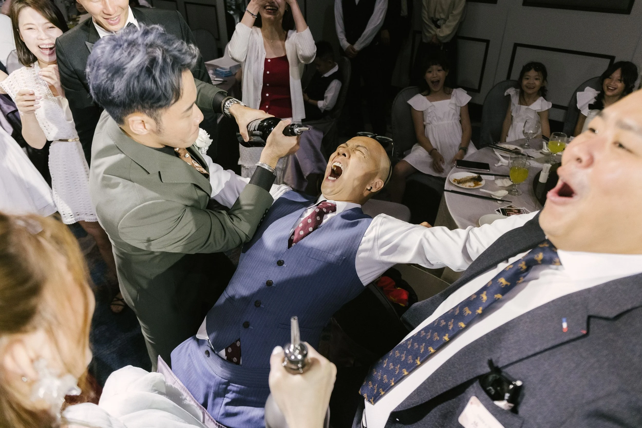 Group of people celebrating at a party, with a man in a blue vest and tie laughing while a person in a suit sprays him with a drink.