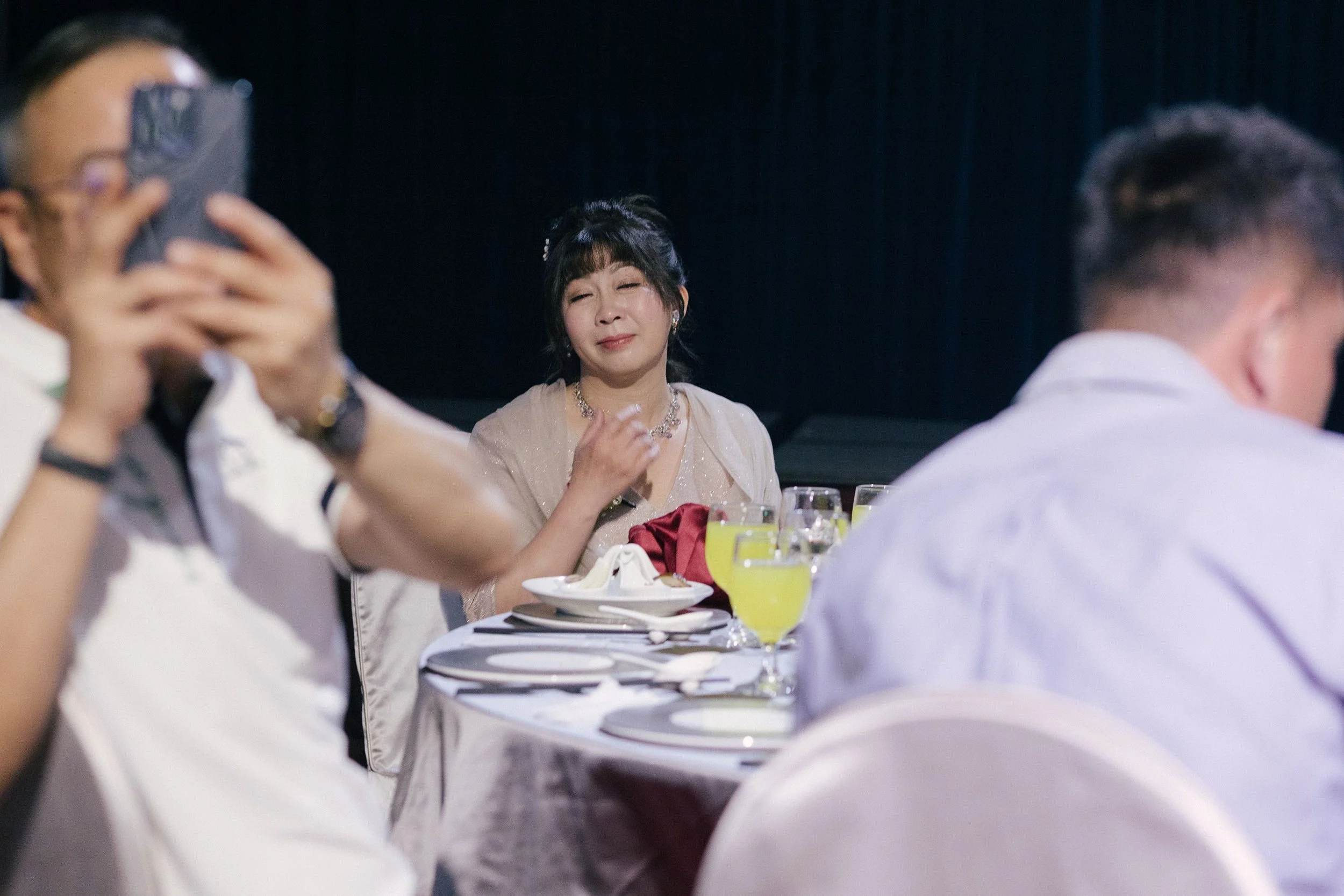 Woman in a beige dress with jewelry smiling at a formal event, seated at a table with wine and food, surrounded by other guests.