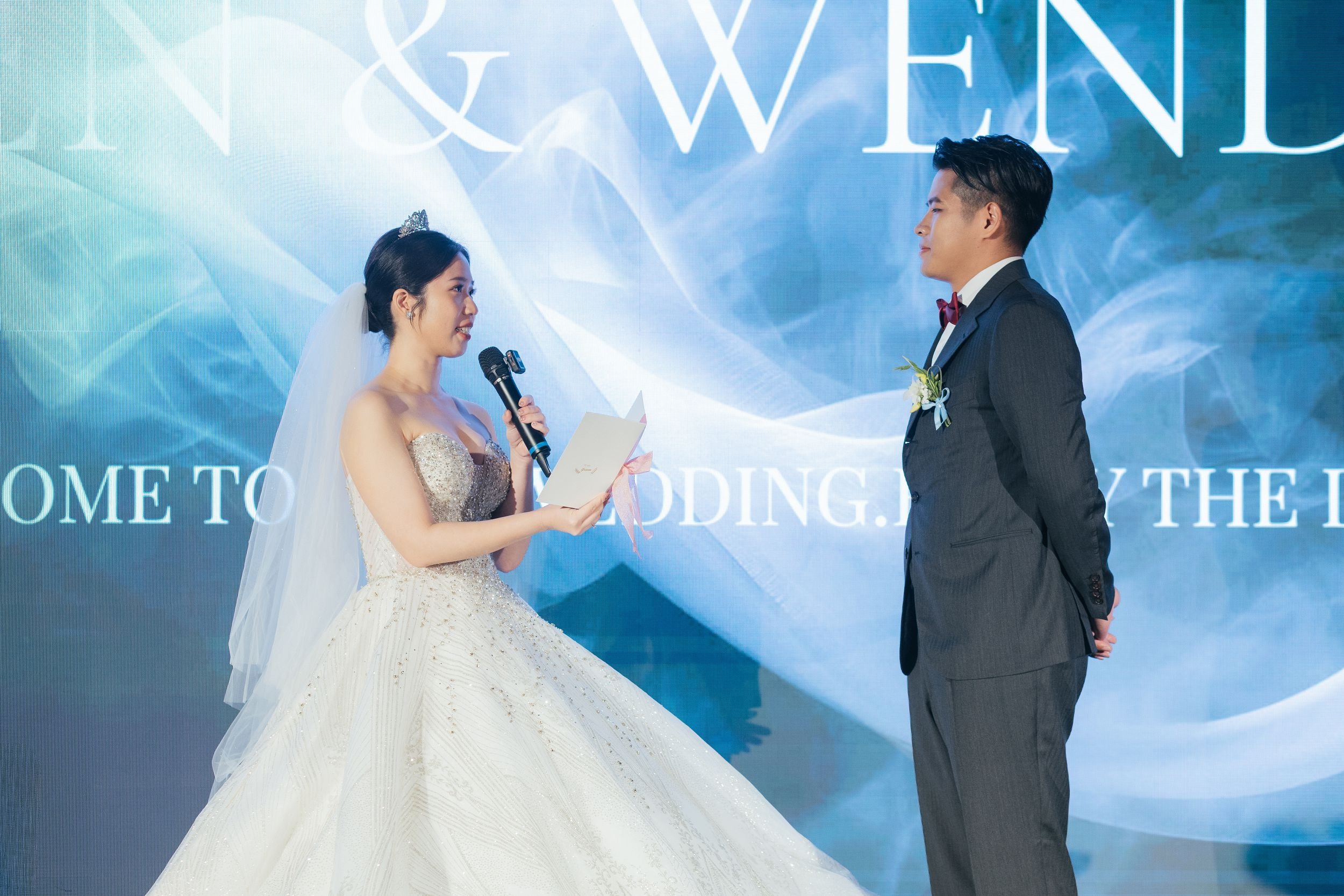 A bride and groom facing each other during their wedding ceremony. The bride is holding a microphone and a paper, wearing a white wedding gown with a tiara and veil. The groom is dressed in a dark suit with a white shirt and red bow tie, with a bouto