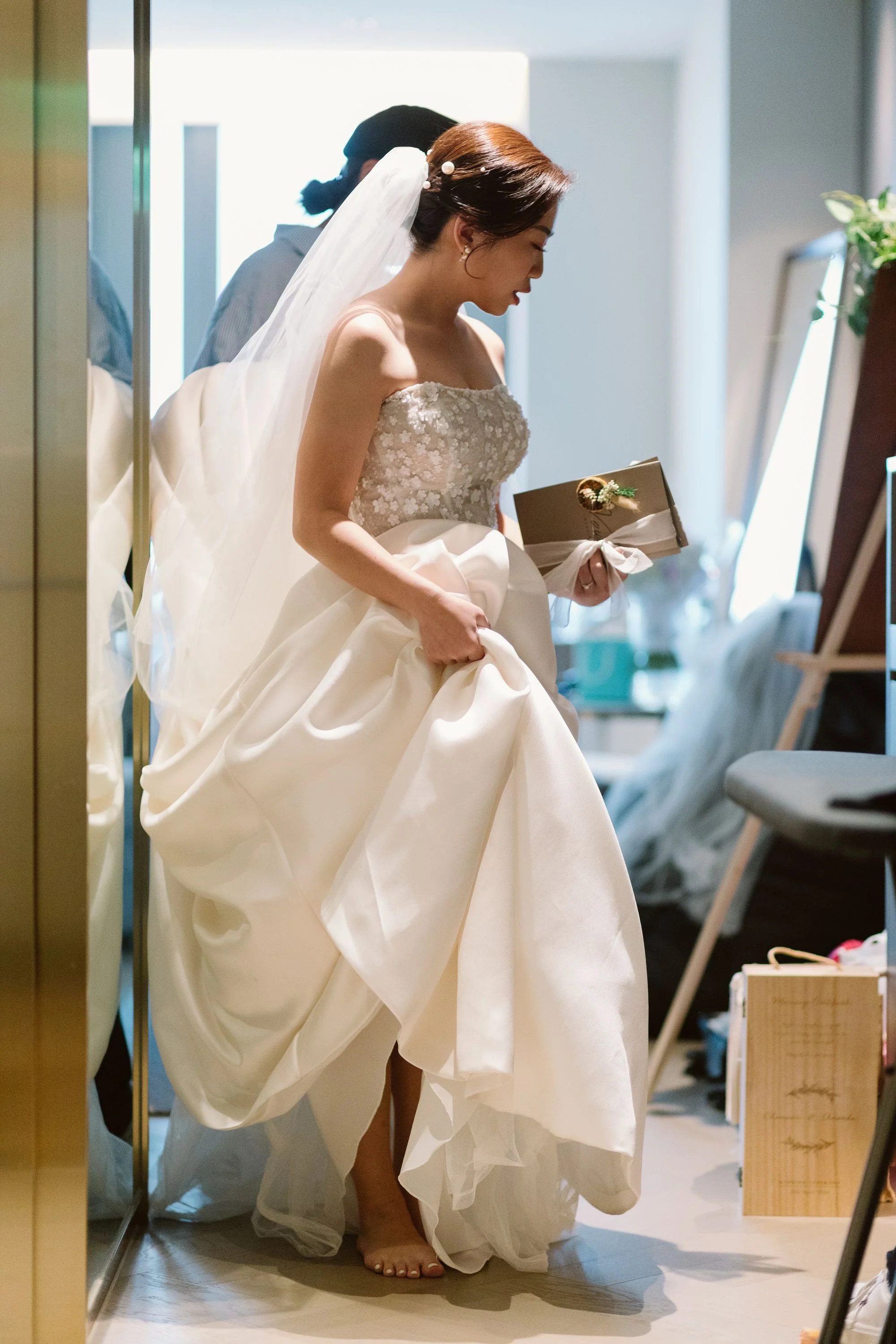 A bride in a strapless wedding dress with floral embroidery holds her gown and a small wrapped gift inside a room, with a man in the background.