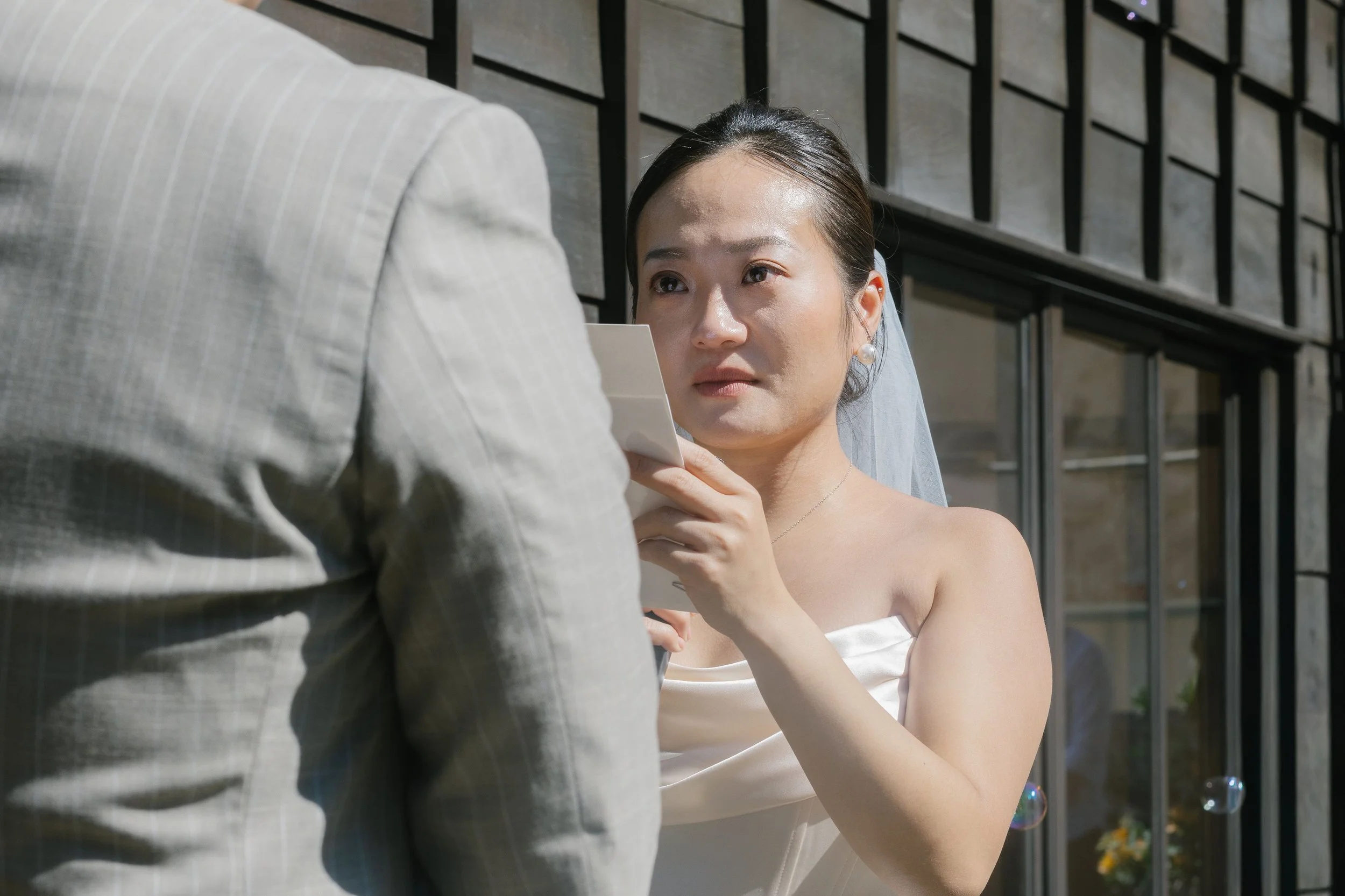 A bride and groom are holding hands outdoors, with the bride looking up at the groom's face. The bride is wearing a strapless wedding dress and pearl earrings, and the groom is dressed in a light-colored suit. They are standing in front of a metal ra