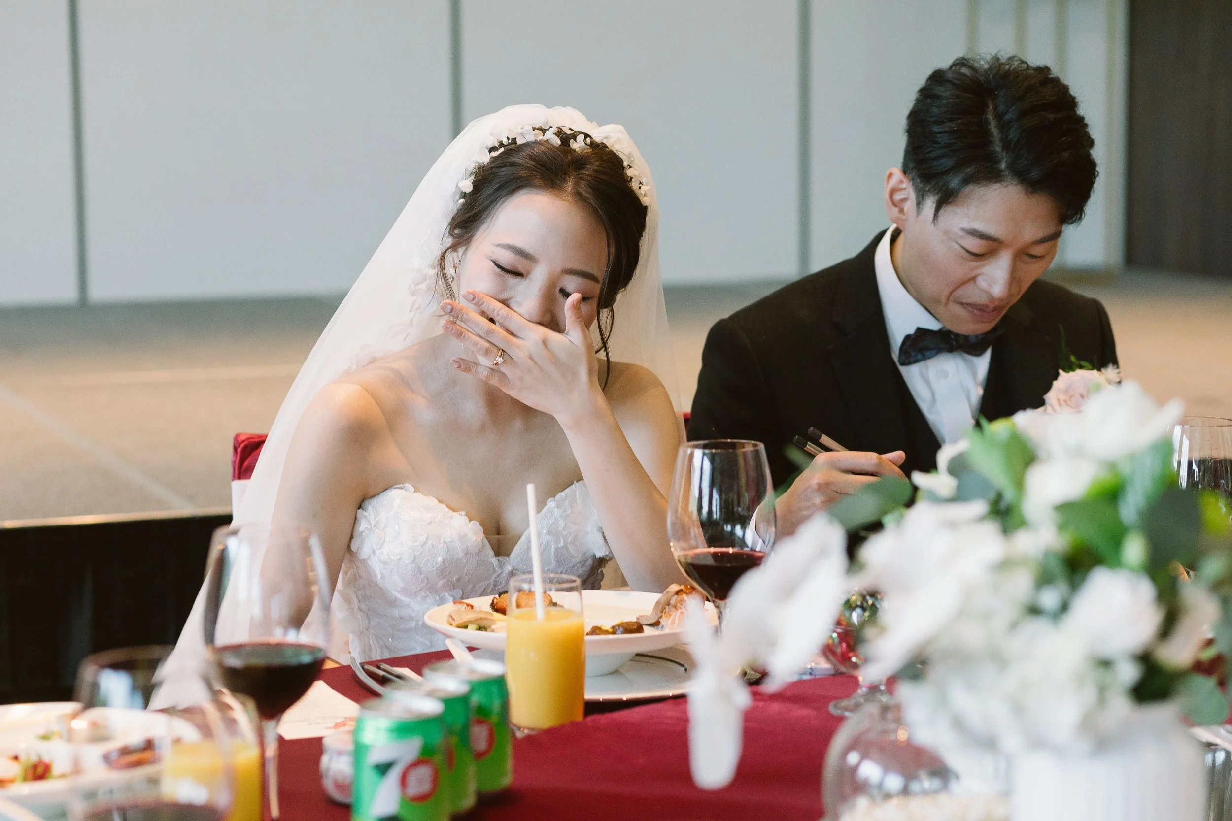 A bride and groom sitting at a table during a wedding reception. The bride is smiling and covering her mouth with her hand, wearing a white dress and veil. The groom is looking down, dressed in a black tuxedo with a bow tie. The table is decorated wi
