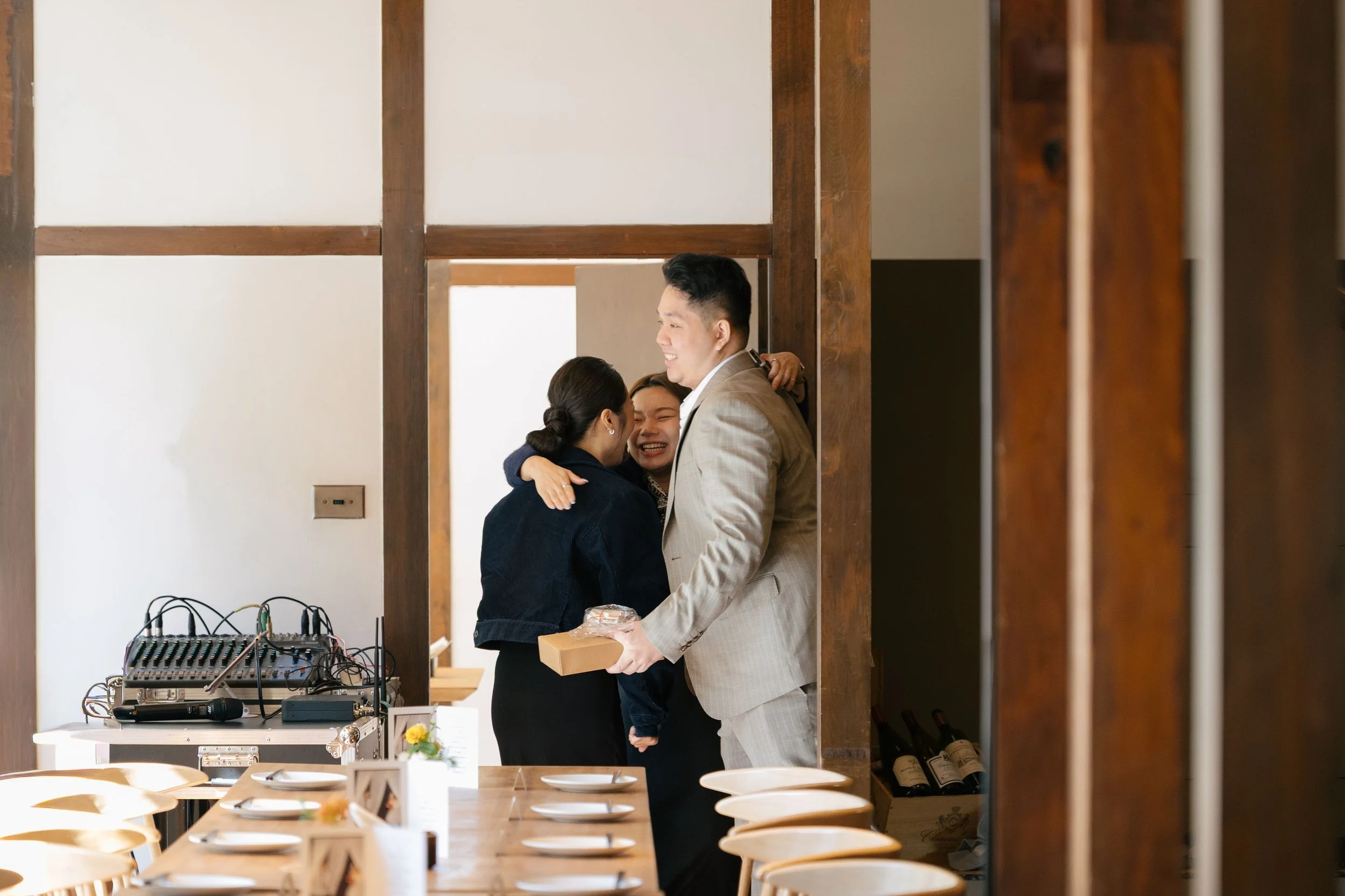 A man in a suit giving a hug and a gift to a smiling woman during a celebration or event in a restaurant or banquet hall.