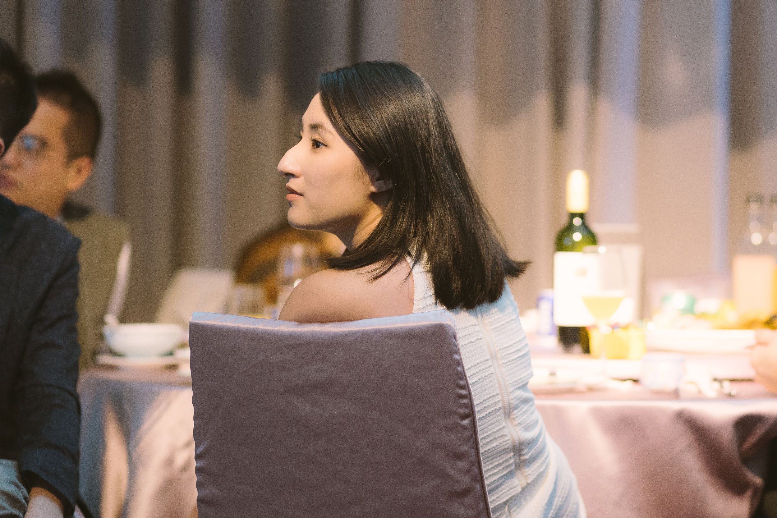 A woman with shoulder-length dark hair and light skin, sitting at a table in an indoor setting, looking to her left. She is wearing a light-colored sleeveless top, and there are bottles and dishes on the table.