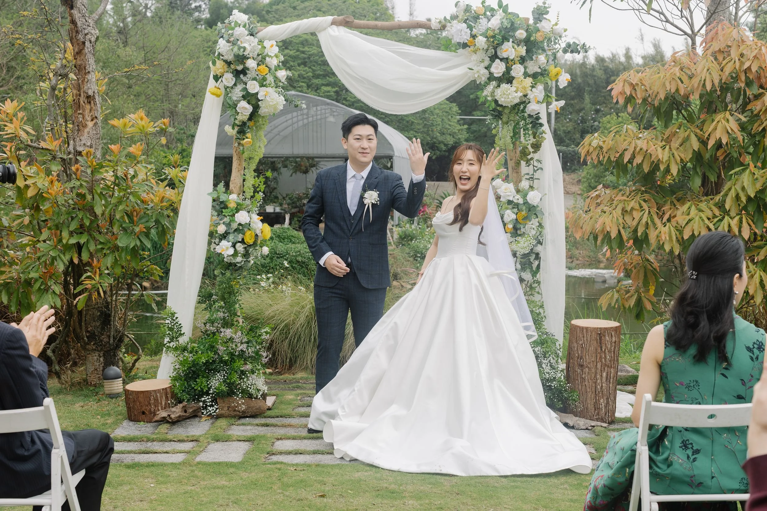 A bride and groom standing under a floral wedding arch outdoors, waving and smiling, as guests sit on white chairs watching them during the wedding ceremony.