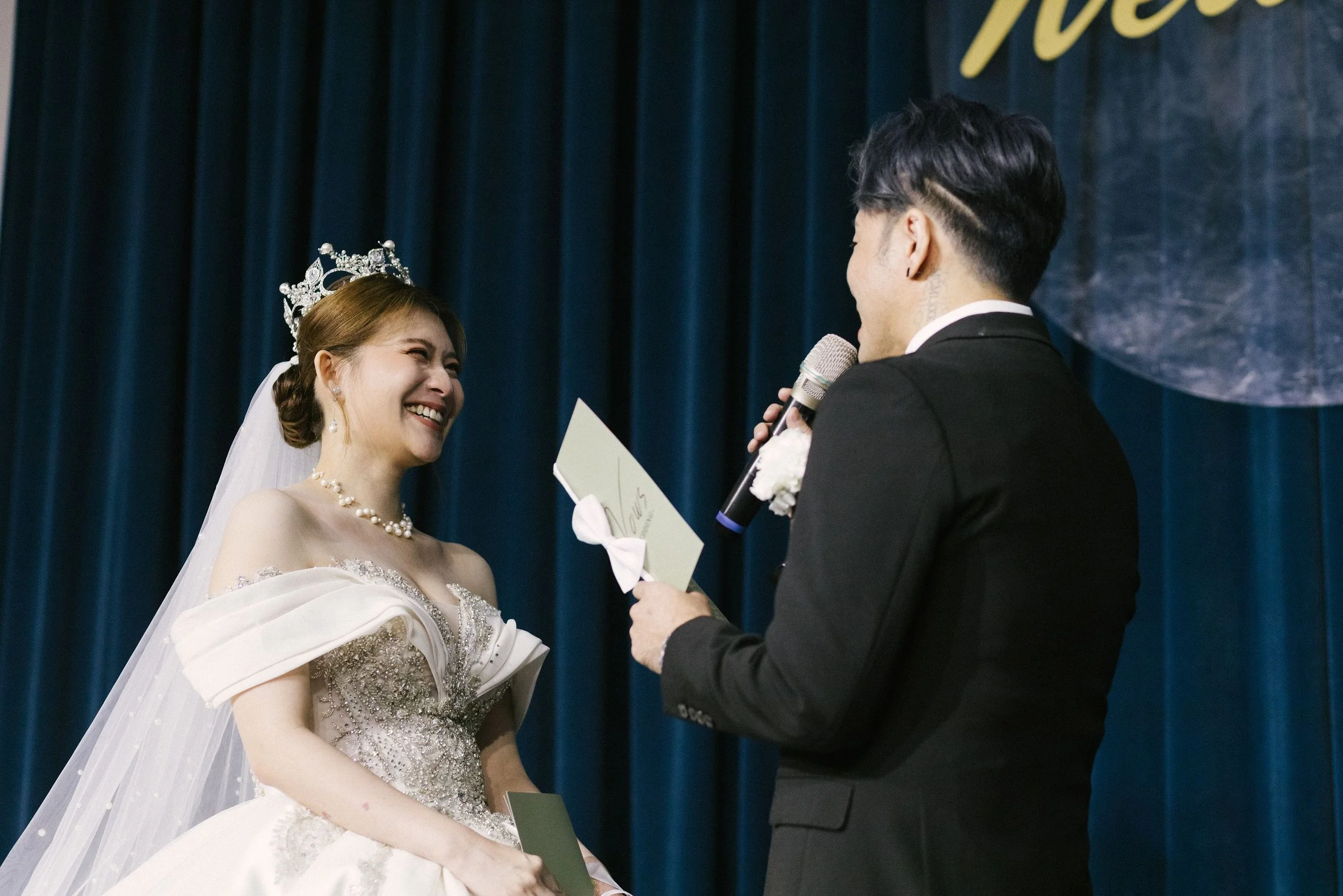 A bride and groom exchanging vows during their wedding ceremony, with the bride smiling joyfully and the groom holding a microphone while reading vows.