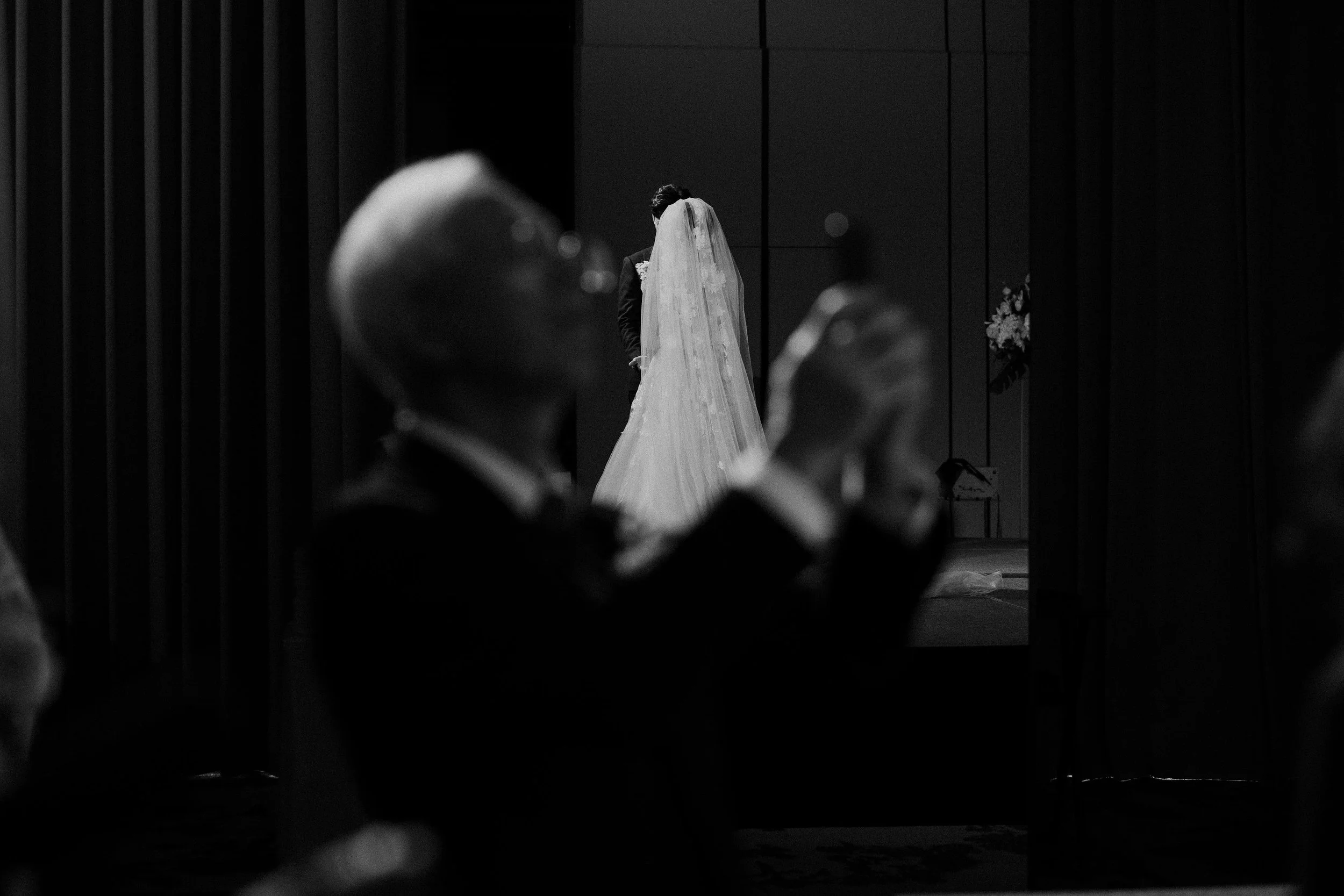 A bride and groom sharing a kiss at their wedding, seen through a gap with out-of-focus guests in the foreground, in black and white.