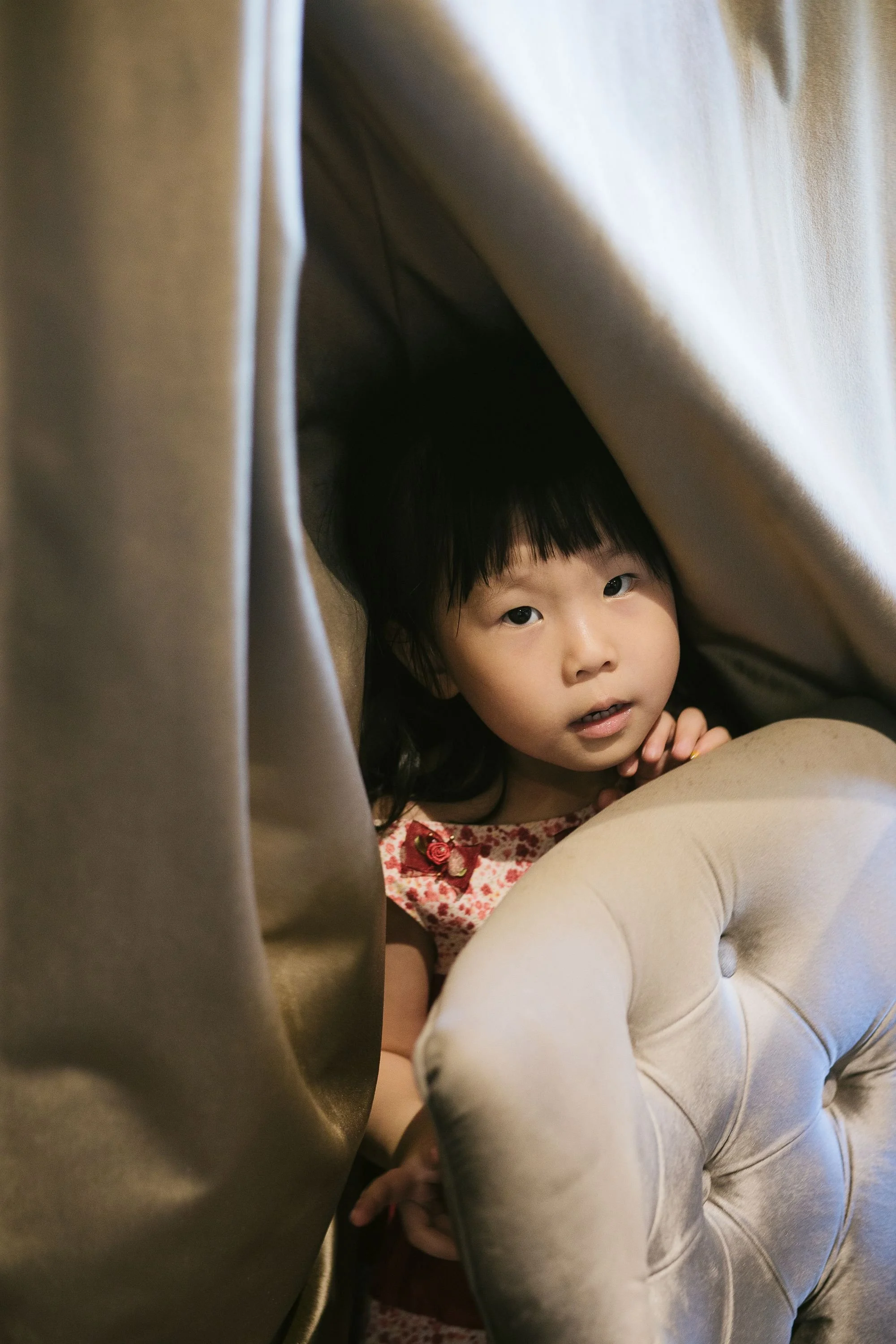 A young Asian girl with dark hair is hiding beneath a blanket or curtain, peeking out and looking toward the camera, with a curious expression. She is lying on top of an adult's lap, wearing a white dress with red floral patterns.