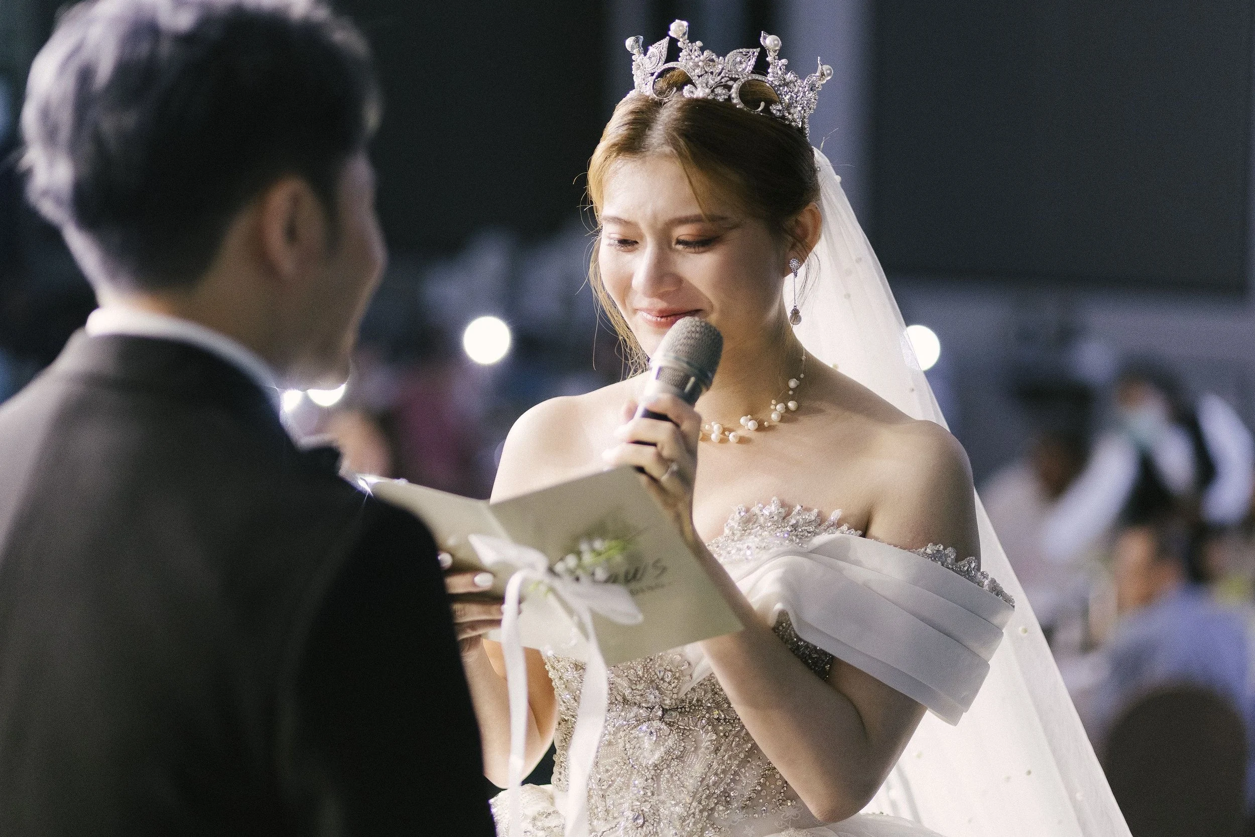 Bride with a tiara and pearl necklace holding a microphone during her wedding ceremony, standing in front of a groom in a dark suit.