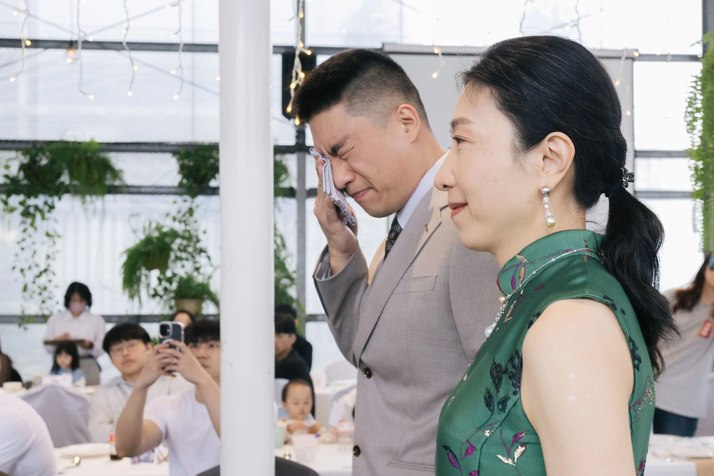 A man in a beige suit and a woman in a green dress are standing close together indoors with string lights above. The man appears to be emotional or crying, and the woman is smiling softly. There are people in the background, some taking photos or loo