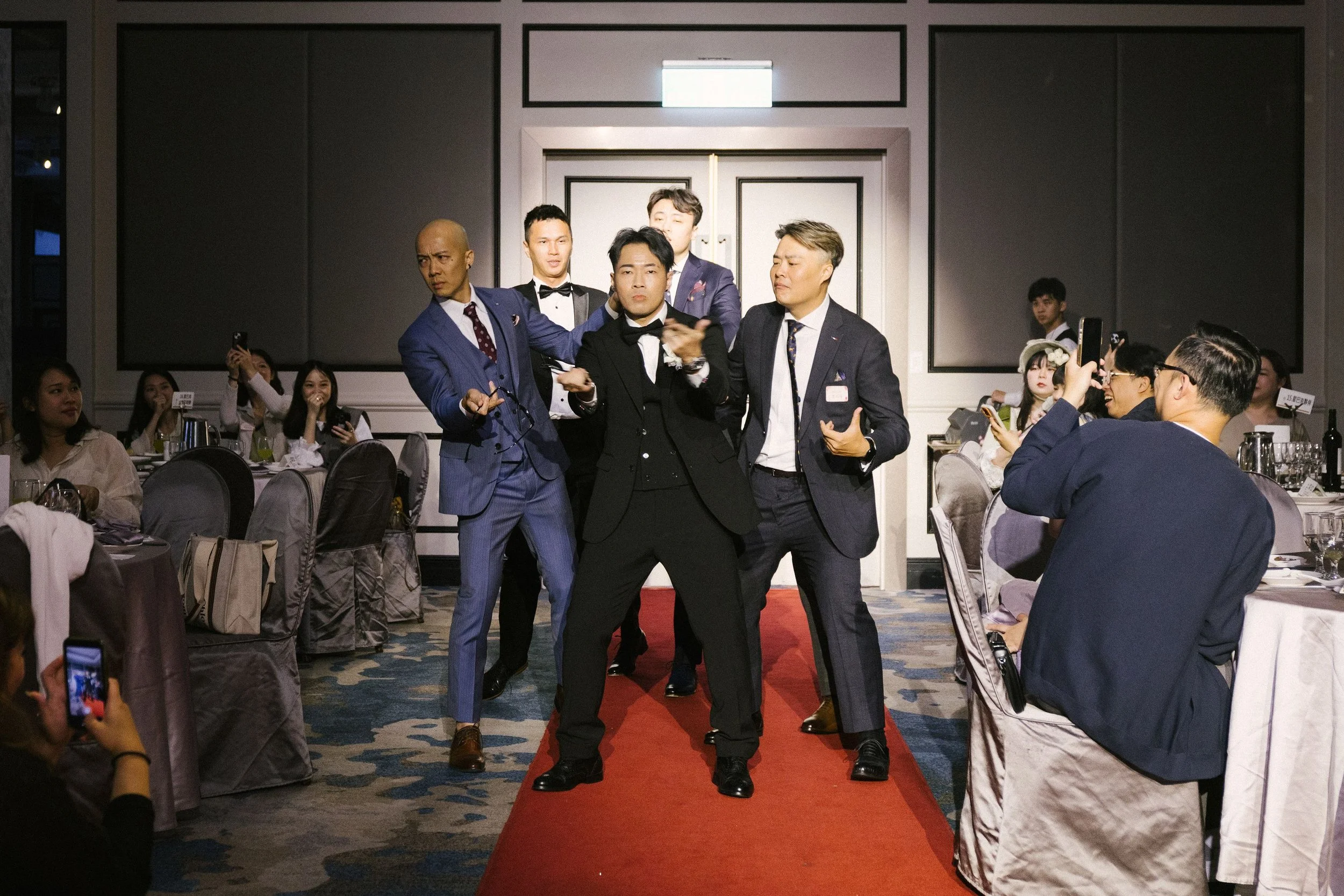 Five men in suits walking down a red carpet at a formal event, with seated guests on each side taking photos and videos.