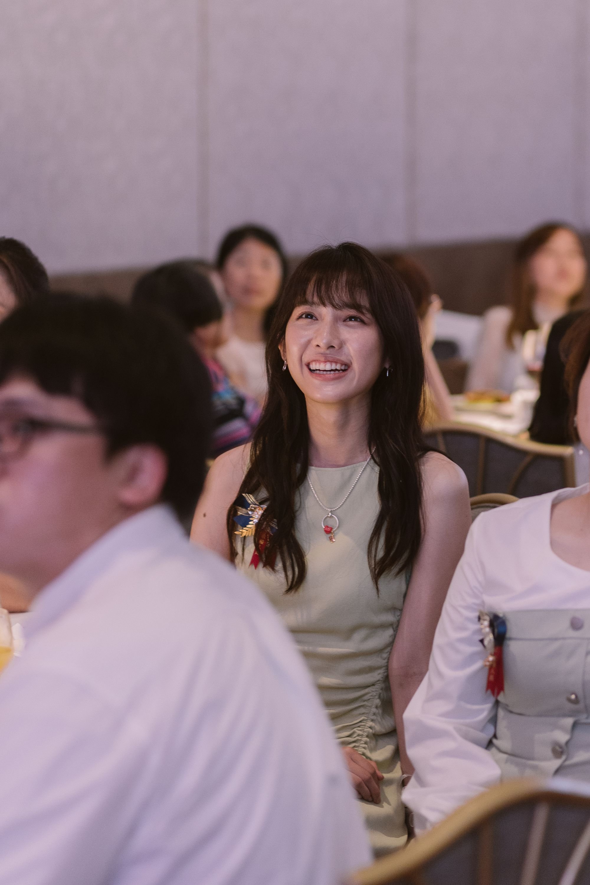 A woman with long black hair and a beige dress with floral accents, smiling at a formal event surrounded by other attendees.