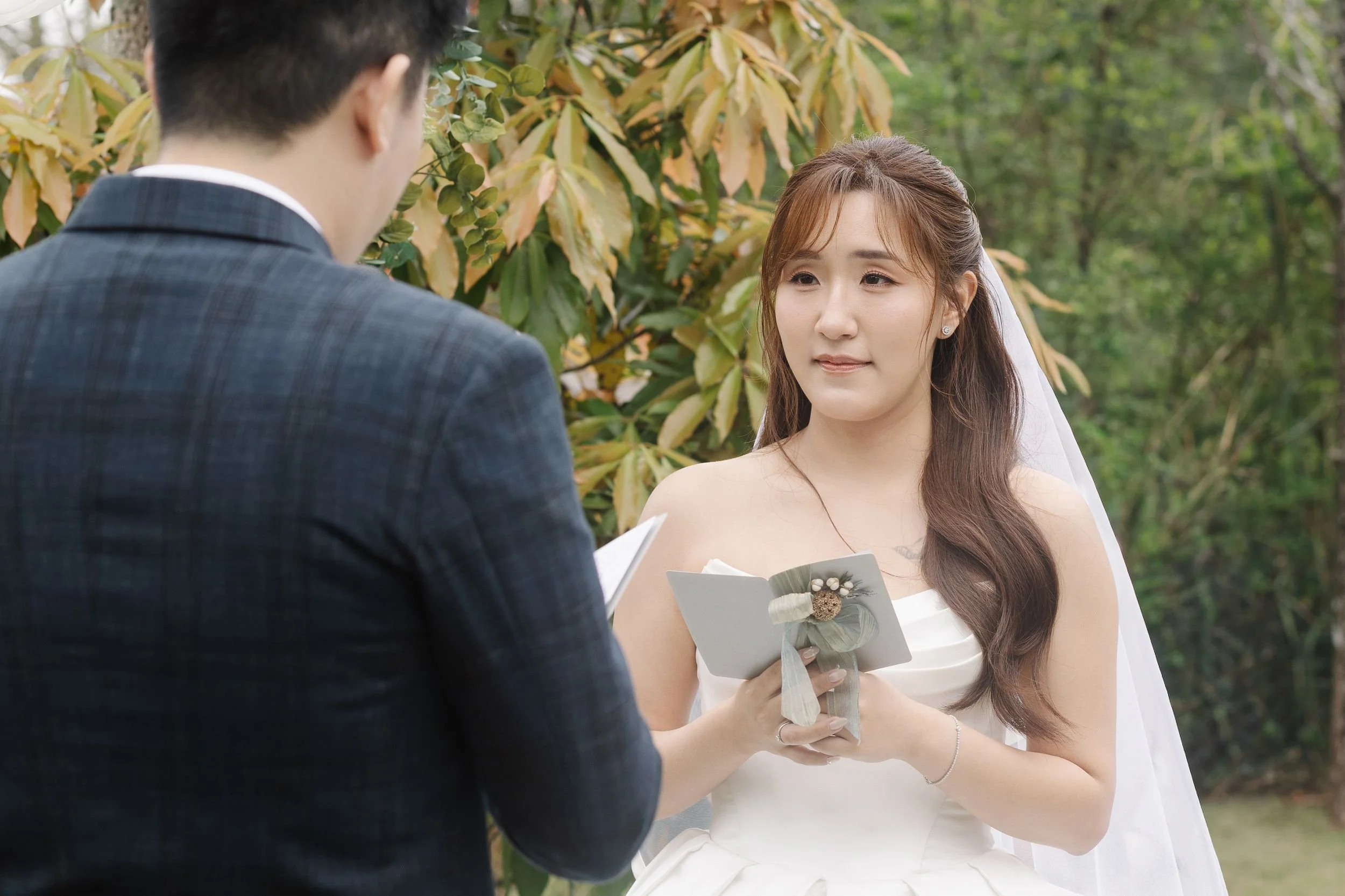 A young woman with brown hair and a white dress is holding a booklet and a small flower bouquet while looking at a man in a suit during an outdoor wedding ceremony in a garden with green foliage.
