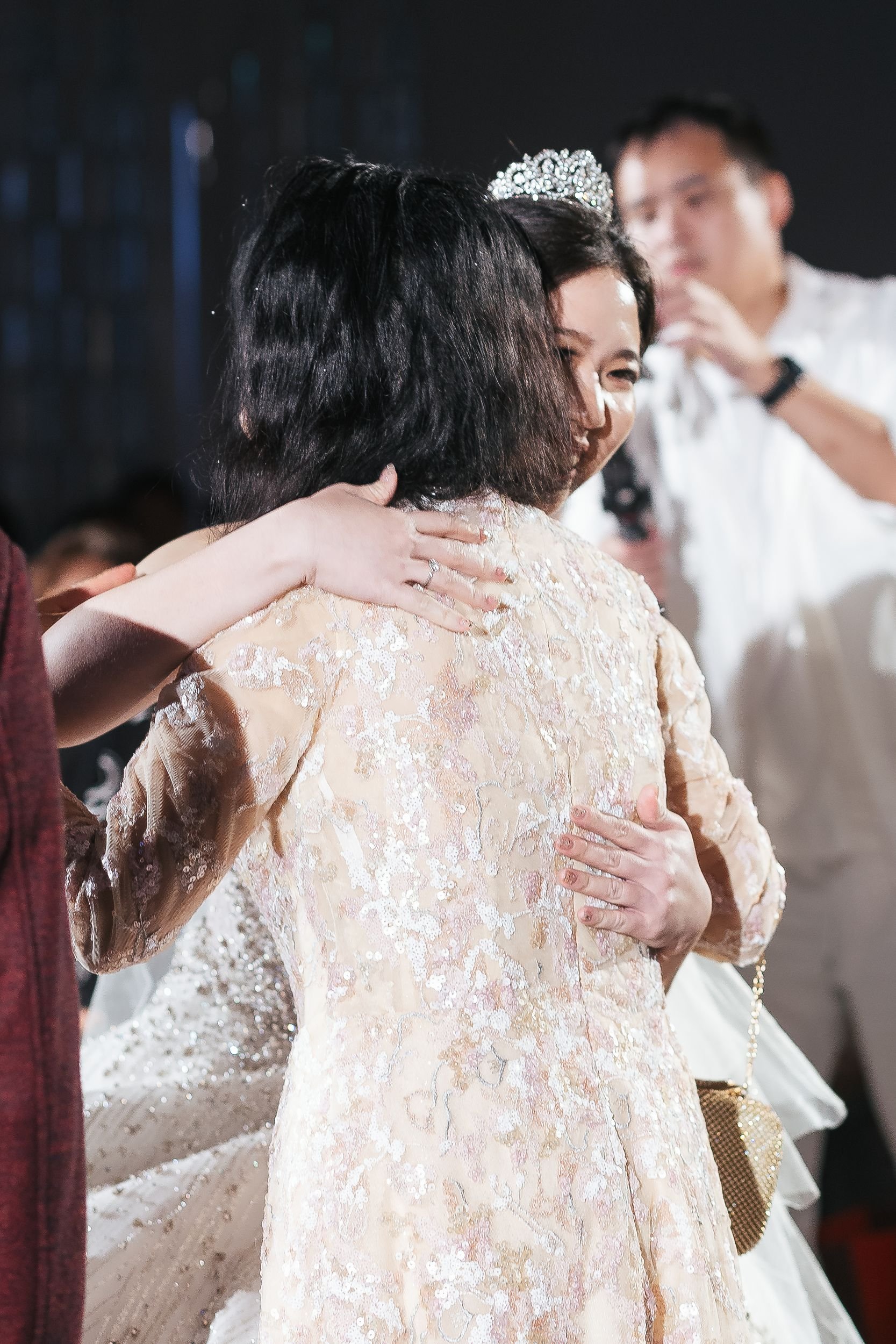 Two women embracing, one wearing a diamond tiara and an elegant dress, with a man in the background, at a celebration or wedding event.