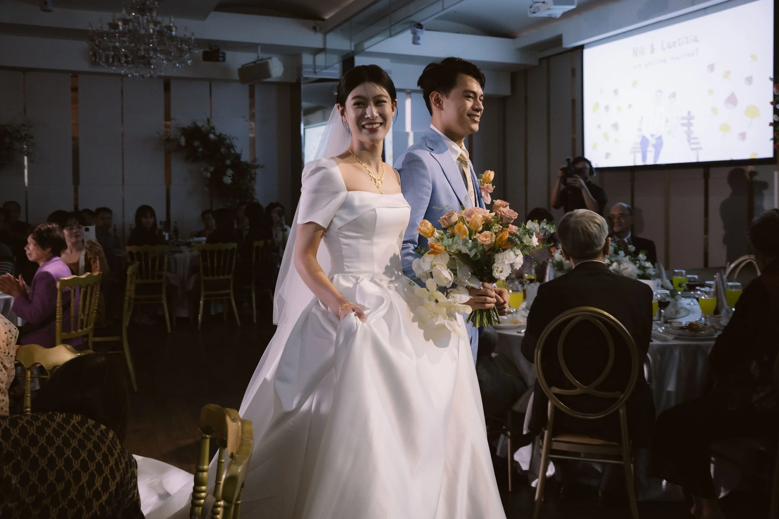 A bride and groom smiling at a wedding reception, standing in front of guests seated at decorated tables with flowers and drinks, with a large screen overhead.