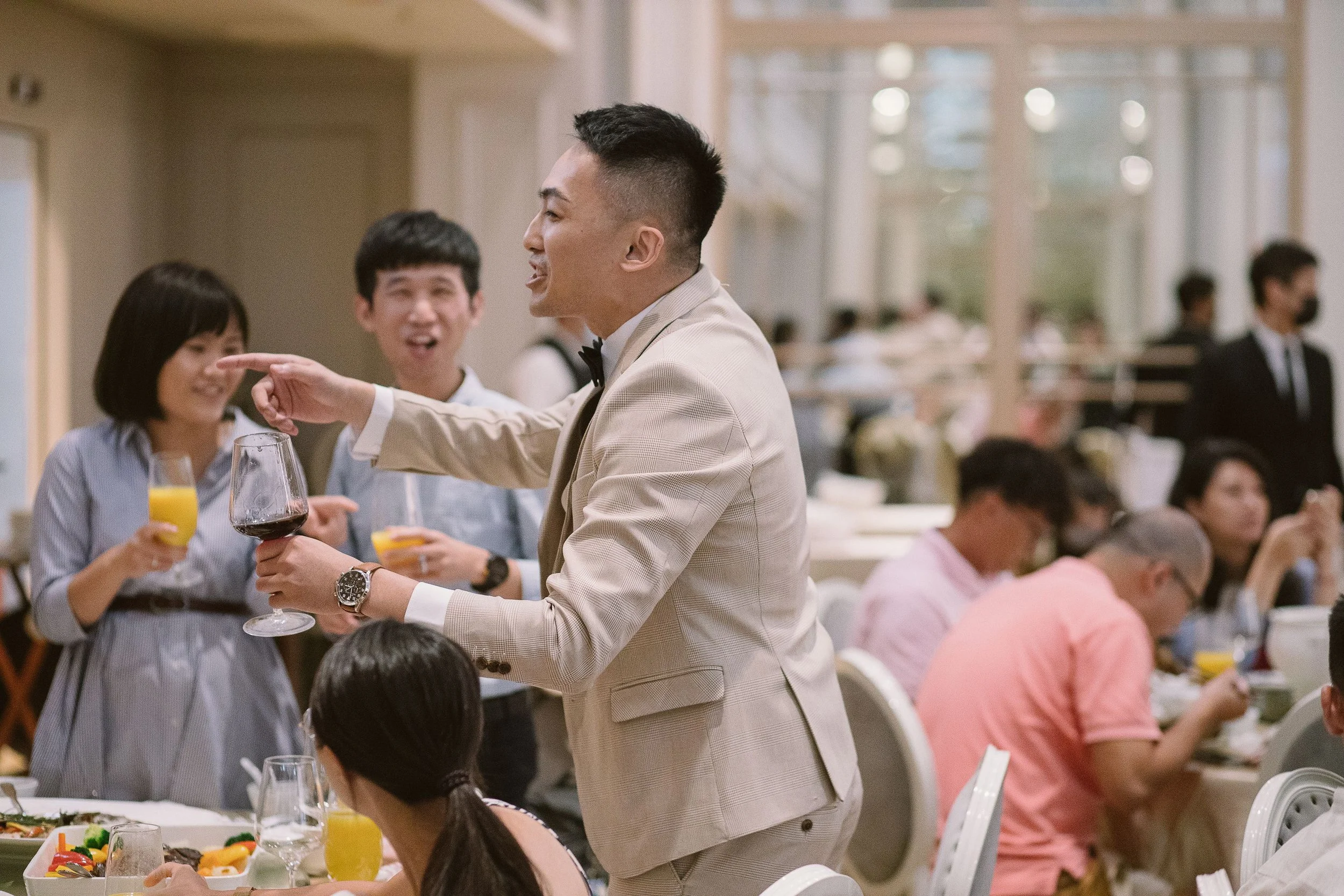 A waiter in a beige suit serving drinks to guests at a formal dining event in a decorated banquet hall.