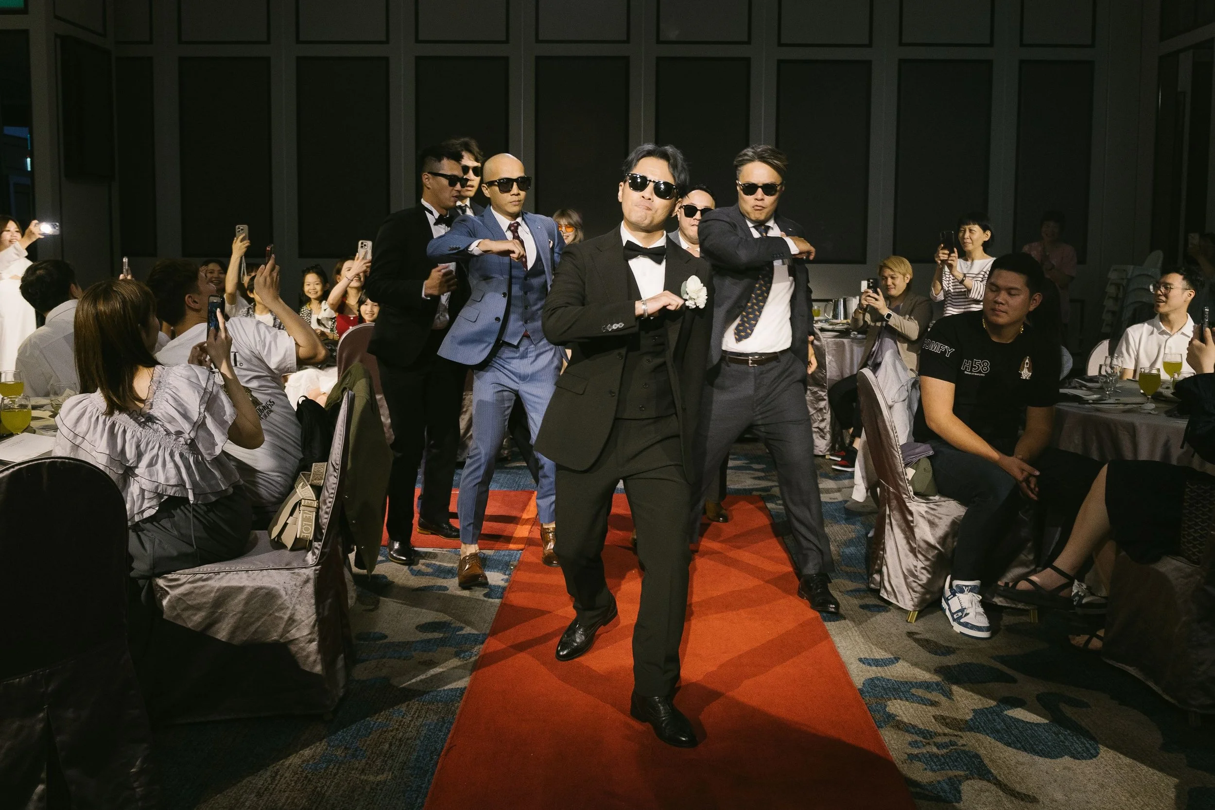 A group of men dressed in tuxedos and sunglasses walking down a red carpet at an event while guests look on and take photos.