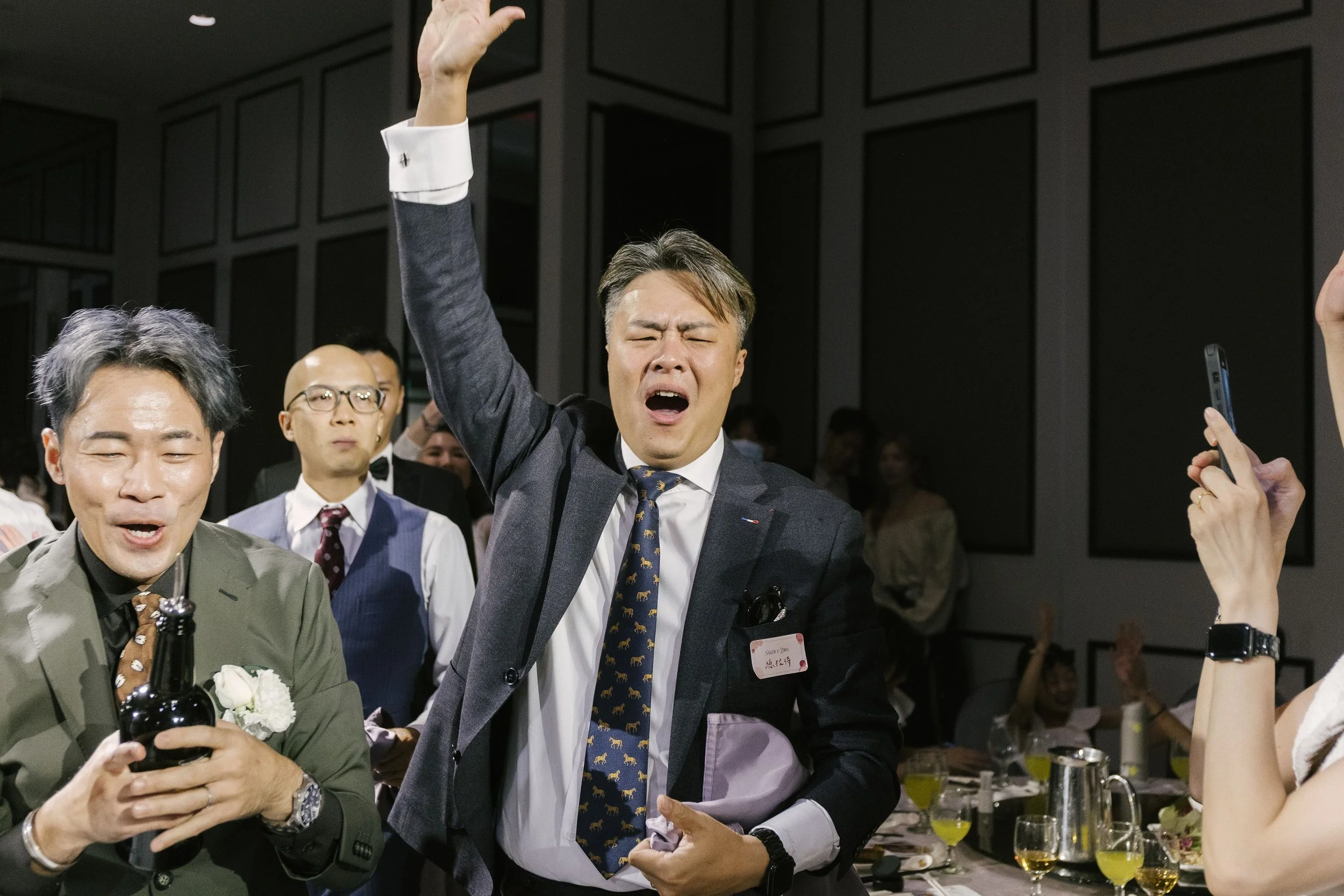 A group of people in formal attire celebrating indoors, with one man in a suit raising his hand and others smiling, holding drinks, in a festive atmosphere.