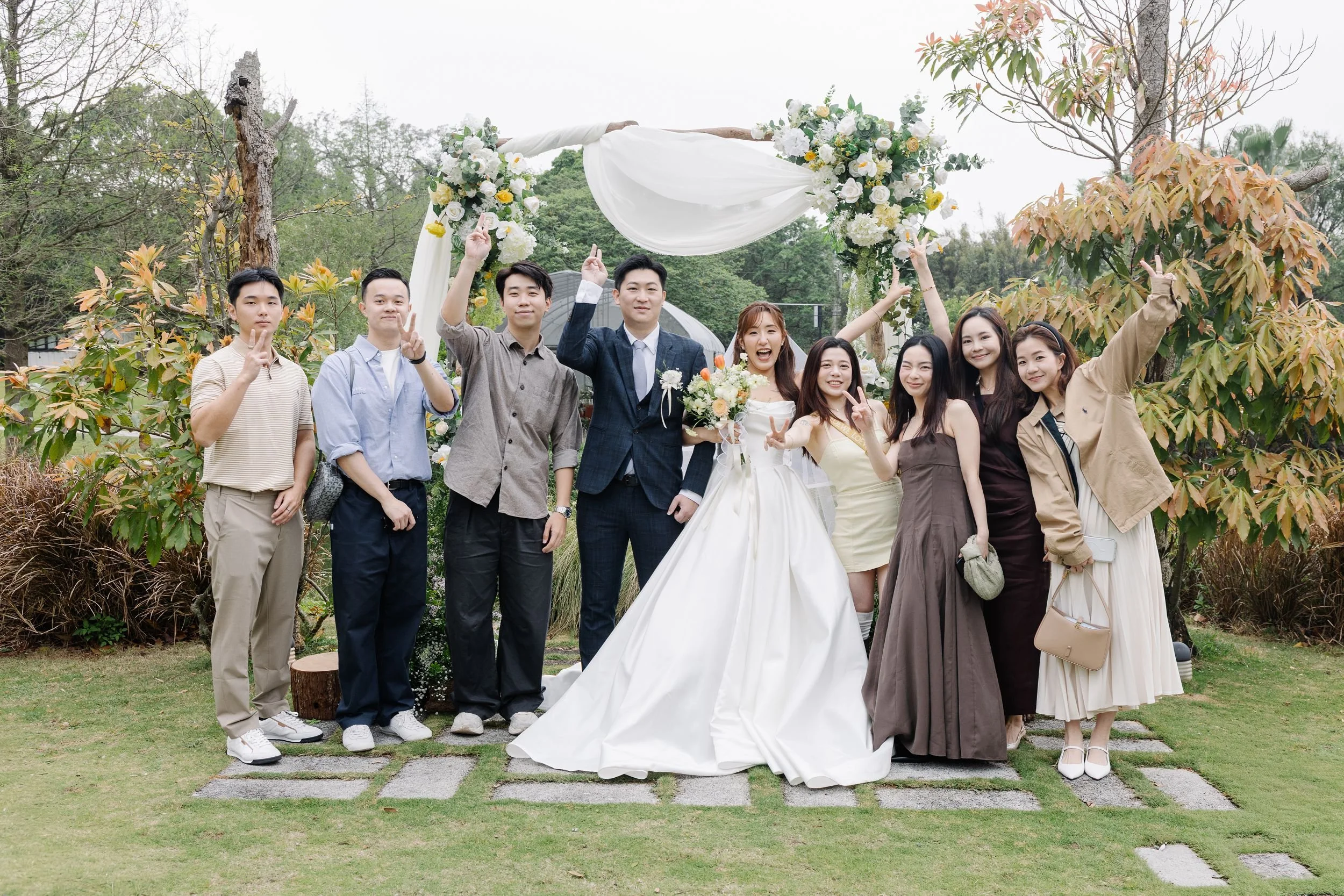 Group of people celebrating wedding outdoors under decorated arch, with some making peace signs and smiling.