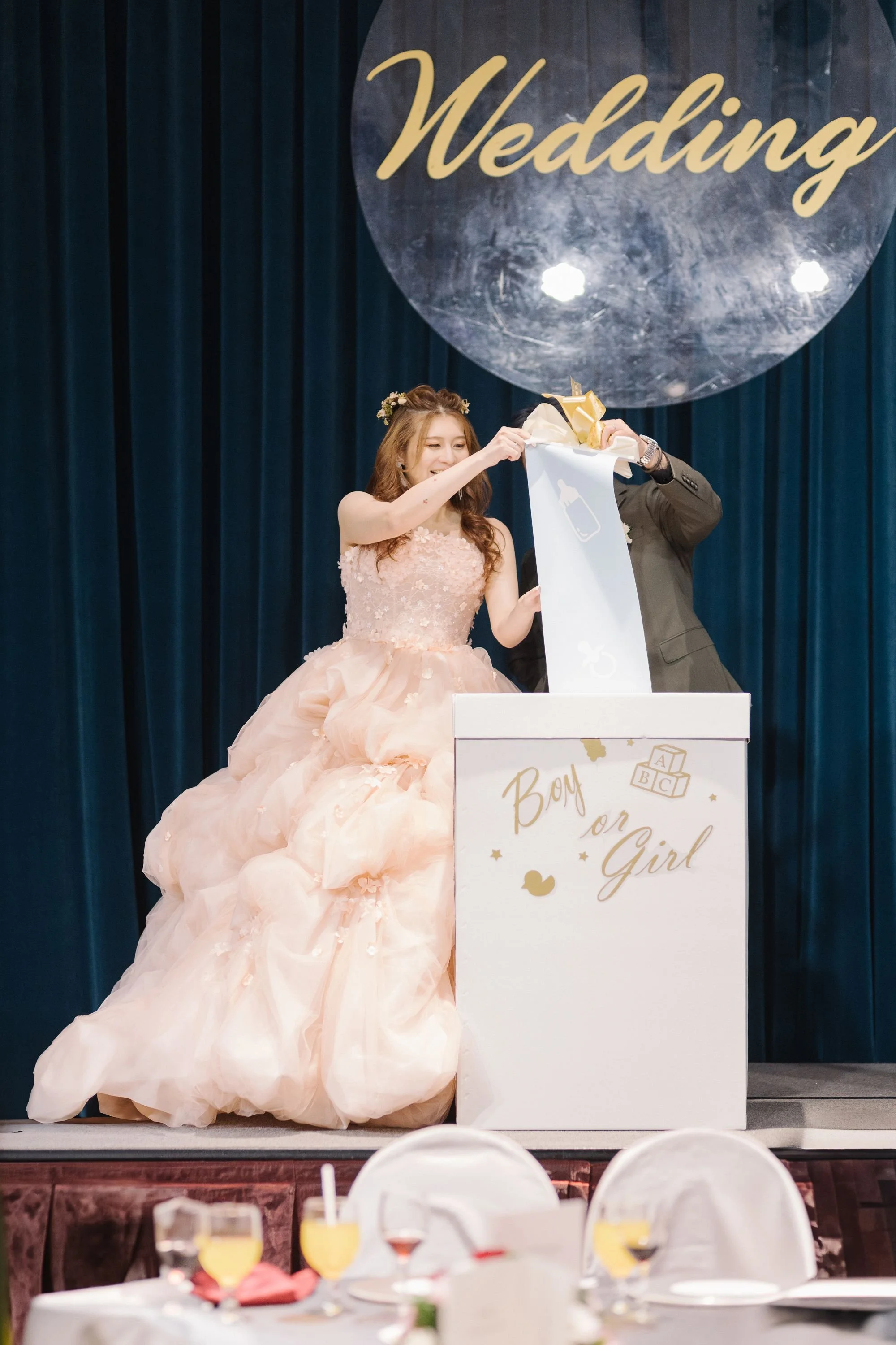 A bride in a pink wedding dress with a floral headband is smiling as she pulls a golden ribbon from a white box on stage, with a sign reading 'Boy or Girl' in front. A man in a suit assists her. The background features blue curtains with a large circ