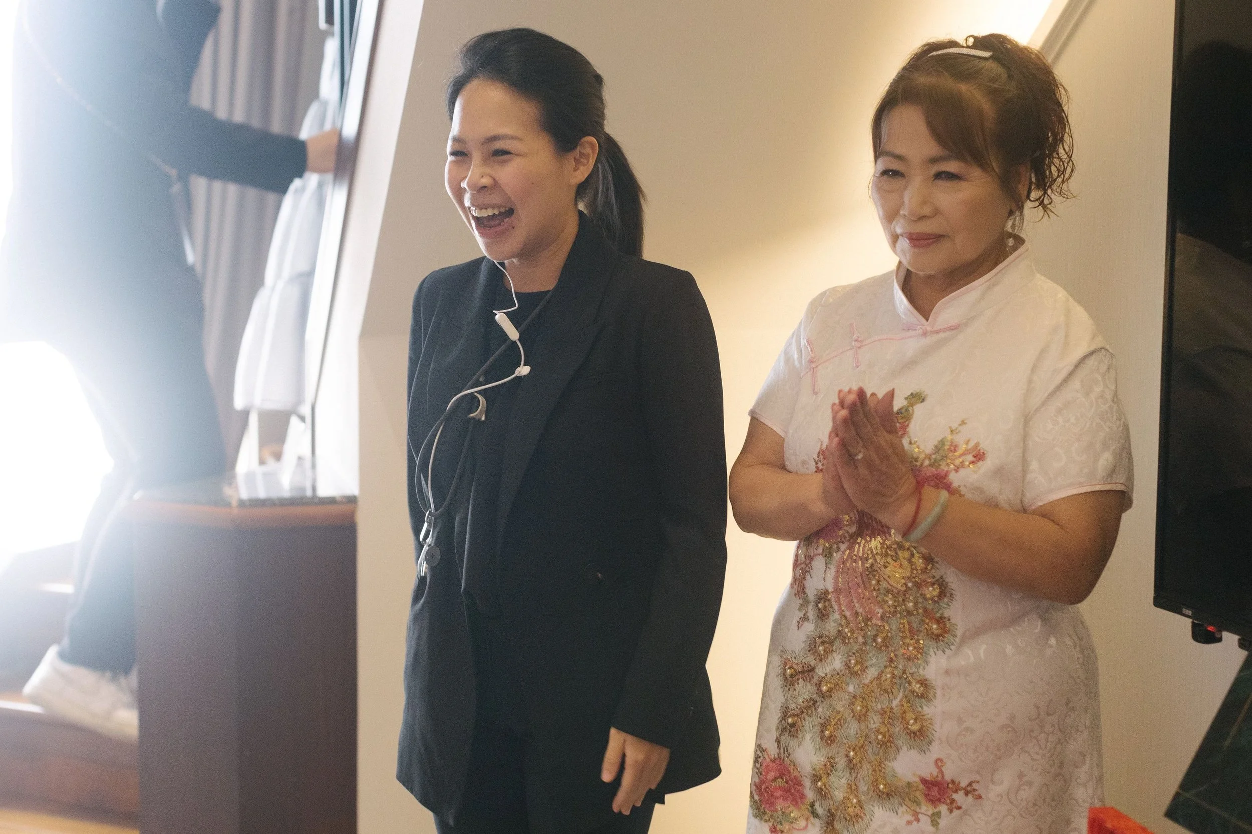 Two women stand side by side, one dressed in black and the other in a traditional white and gold dress, in a room with a warm light. The woman in black is smiling and wearing a headset with a microphone, while the woman in traditional attire has her 