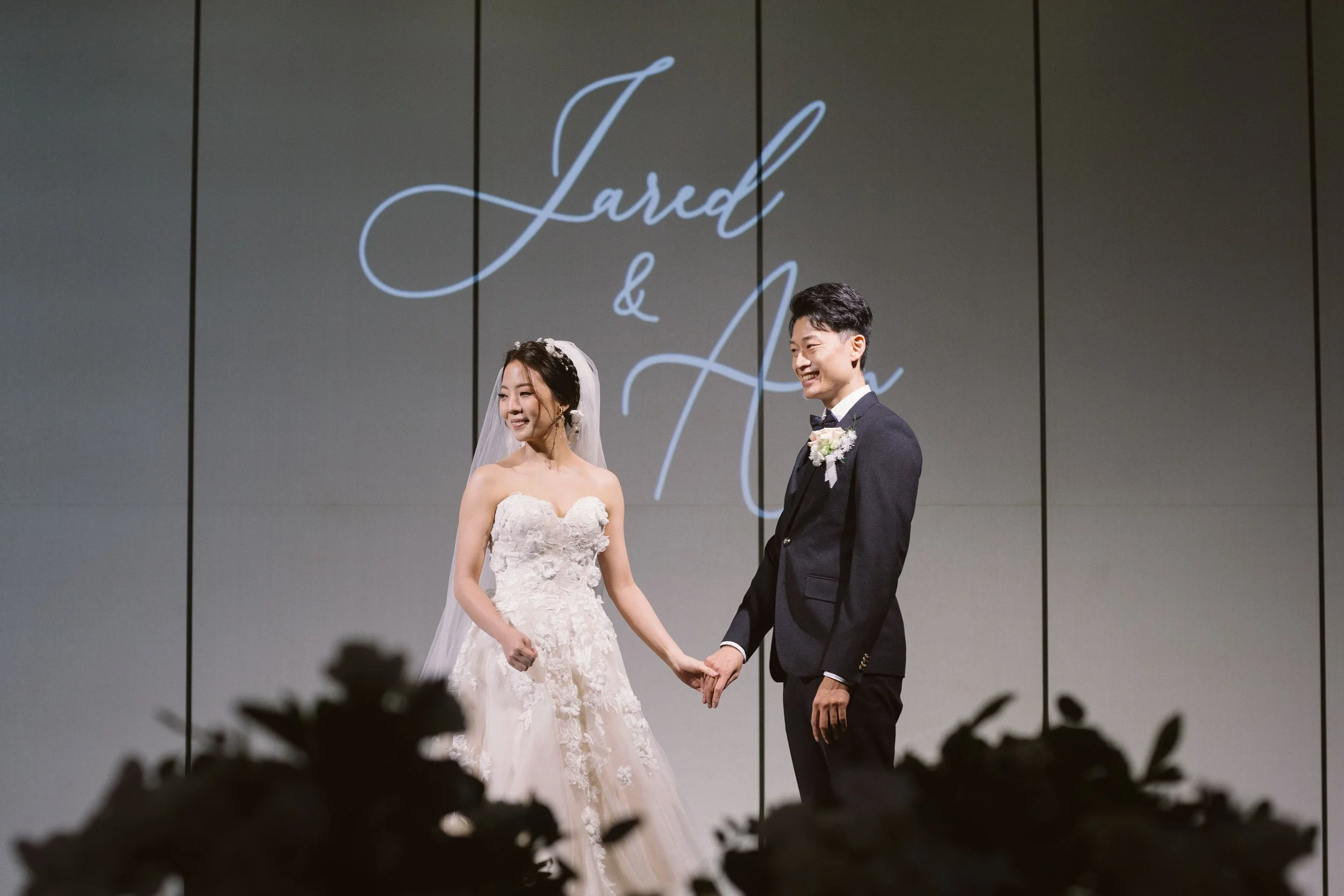 Bride and groom holding hands during wedding ceremony, smiling, with wedding vows written in cursive behind them.
