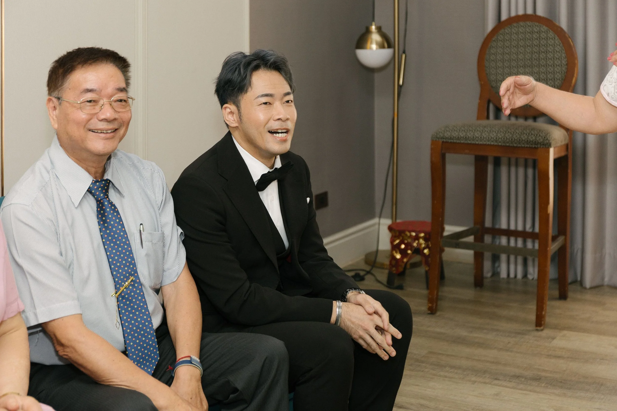 Two men sitting in a row, dressed formally, in a room with a gray wall and a decorative chair behind them. One man is smiling, the other is talking. A person's arm is visible on the right side of the image, gesturing towards the seated men.