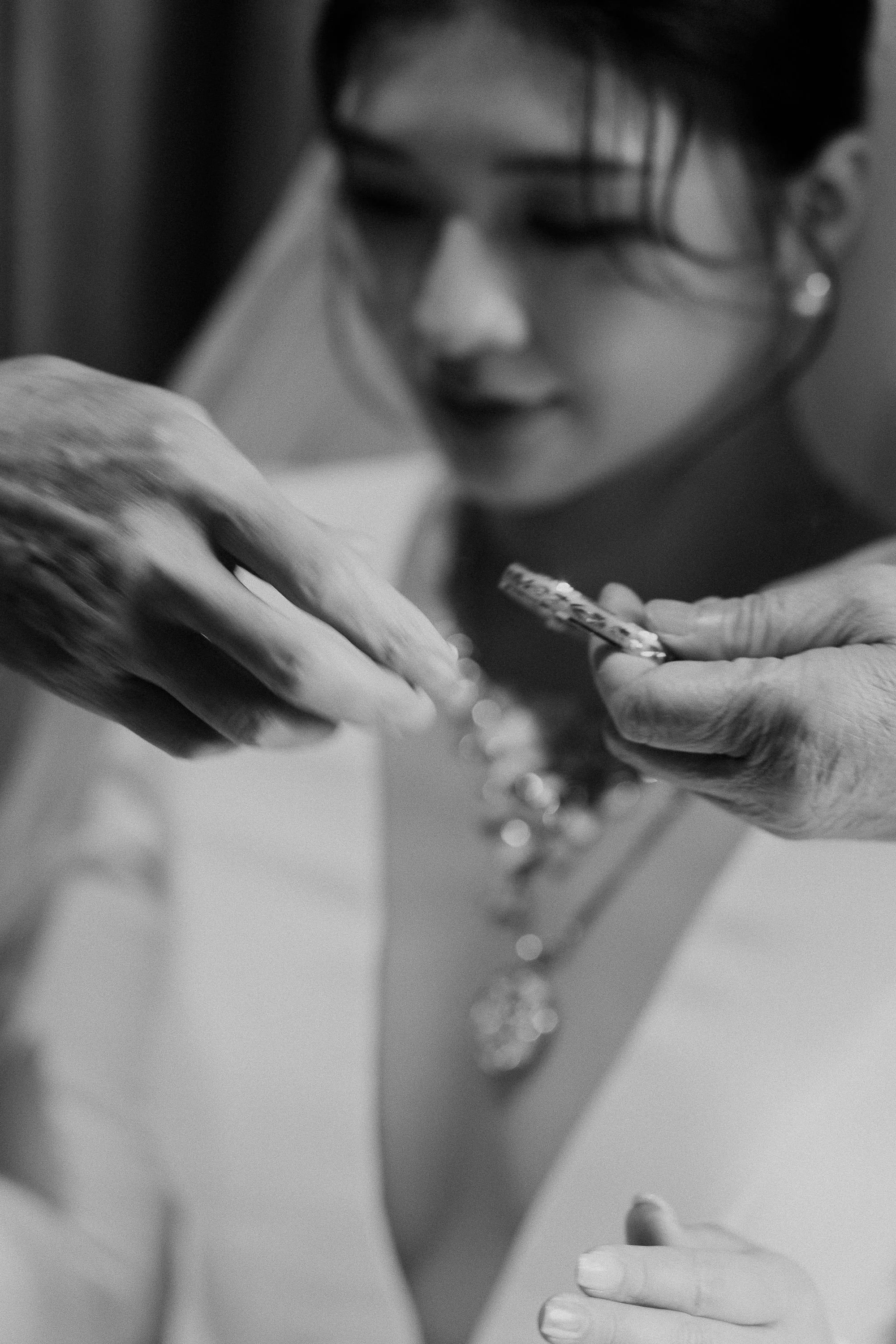 A woman with dark hair and earrings, looking down, holding a small object, with her other hand resting on her chest. The image is in black and white.
