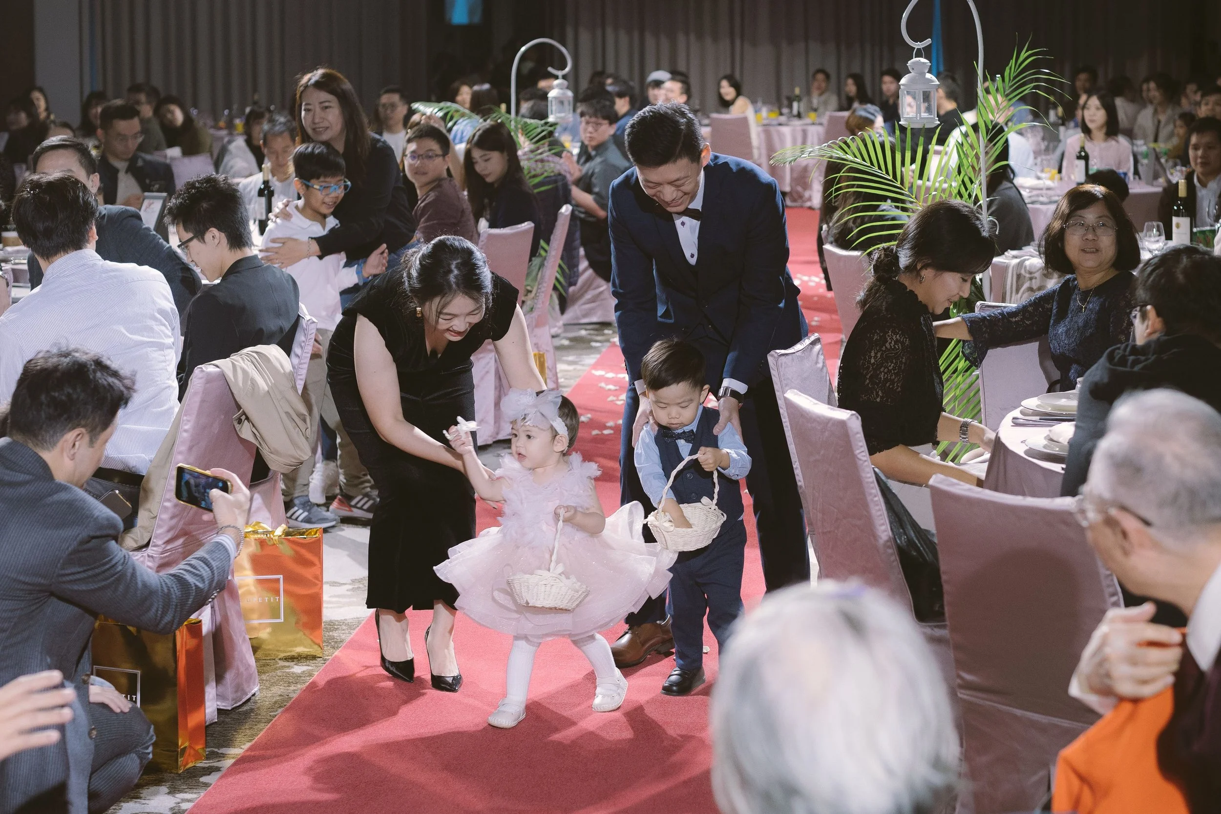 Children in fancy costumes participating in a children's fashion show with adults and spectators watching inside a banquet hall.