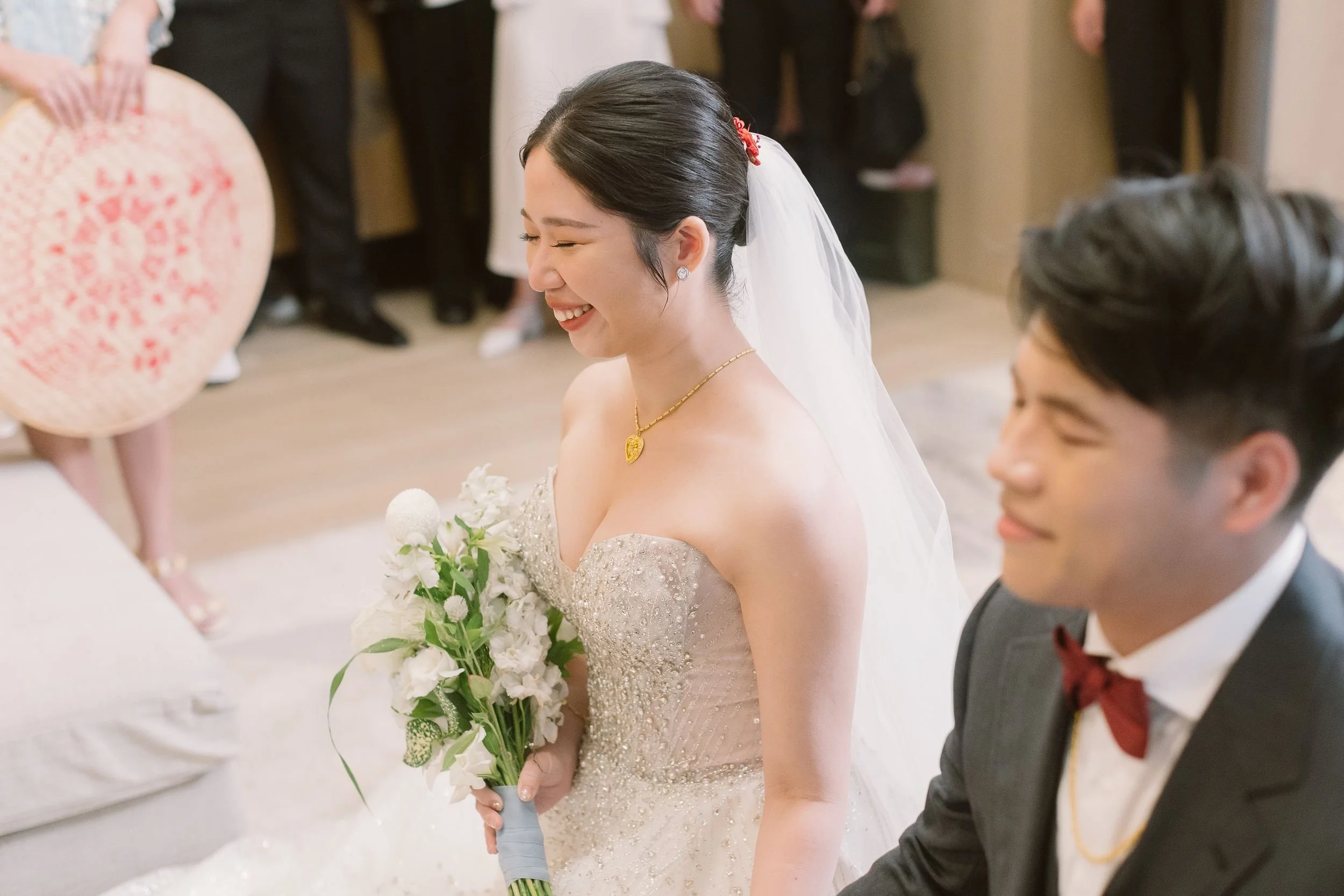 A smiling bride in a wedding gown holding a bouquet of white flowers, standing next to a groom in a dark suit with a red bow tie, during a wedding celebration.