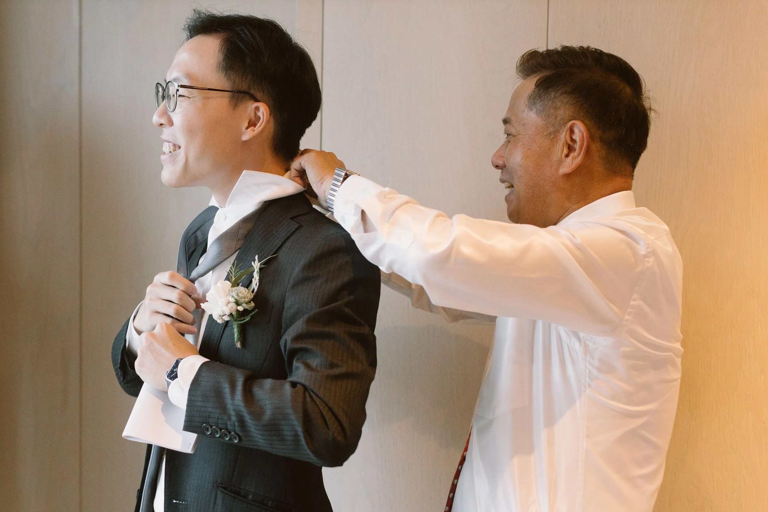 A man in a dark suit, white shirt, and glasses is smiling and holding a paper. Another man, wearing a white shirt, is adjusting the other man's tie.