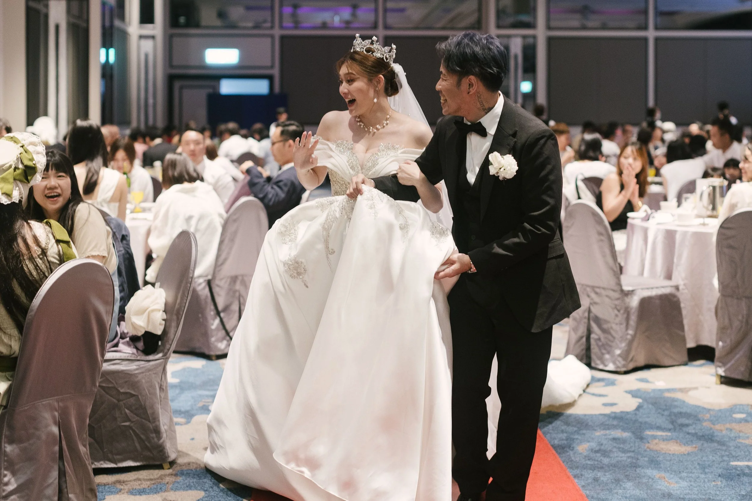 A bride and groom holding hands, smiling, and celebrating at their wedding reception. The bride is wearing a white wedding dress and a tiara, and the groom is in a black tuxedo. Guests are seated at tables in the background.