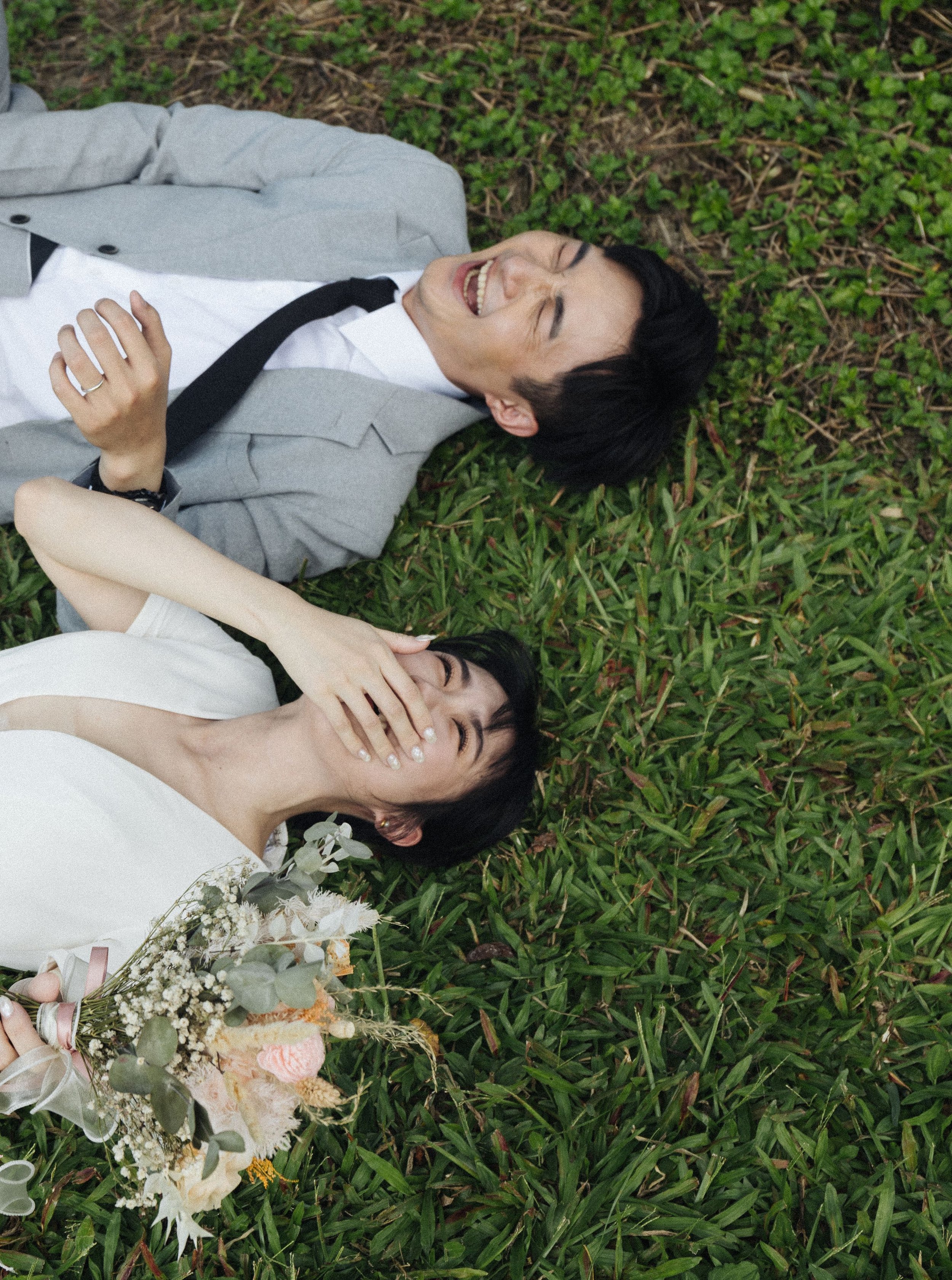 Two women lying on grass, one in a gray blazer and white shirt, the other in a white dress, smiling and covering one another's eyes, with a bouquet of flowers nearby.