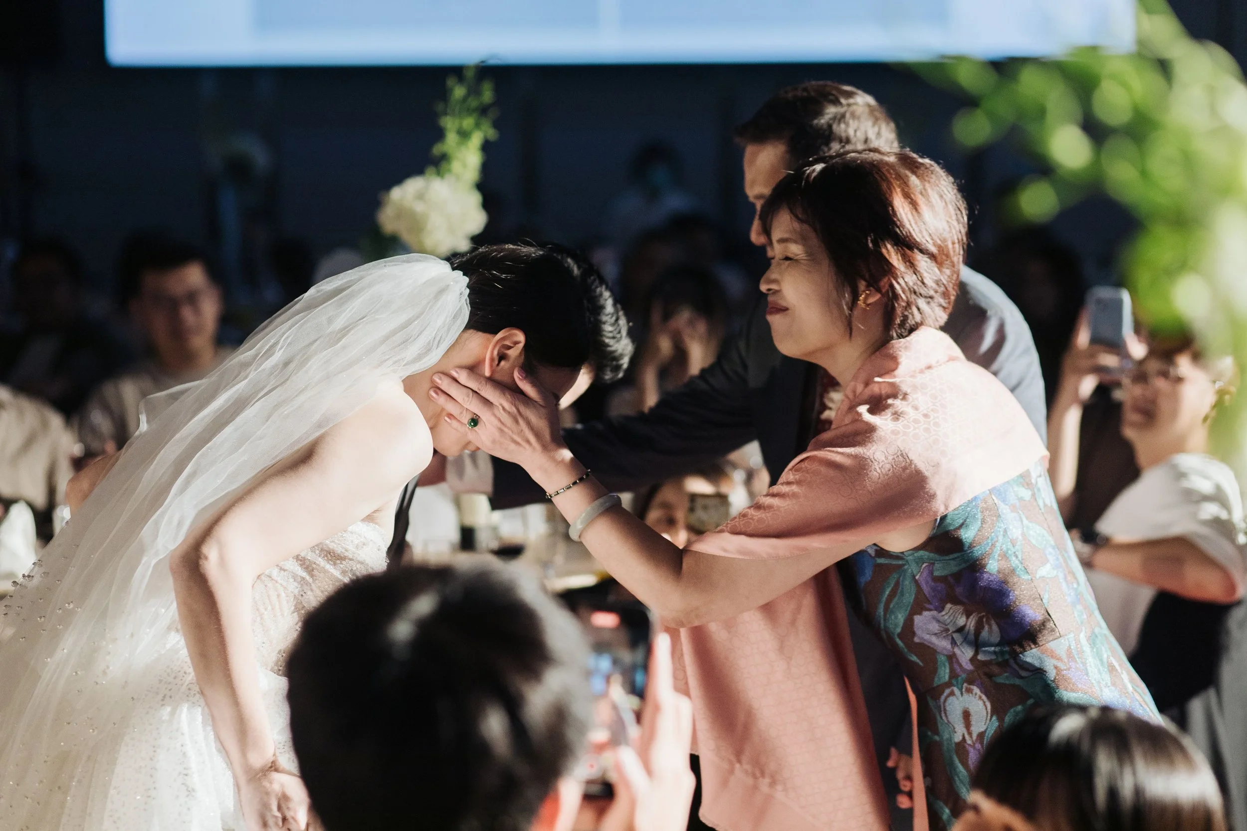 A wedding ceremony with a bride, groom, and a woman, possibly the mother of the bride, participating in a cultural ritual. The woman is holding the bride's face with both hands, and the bride is bowing her head. Guests are visible in the background.