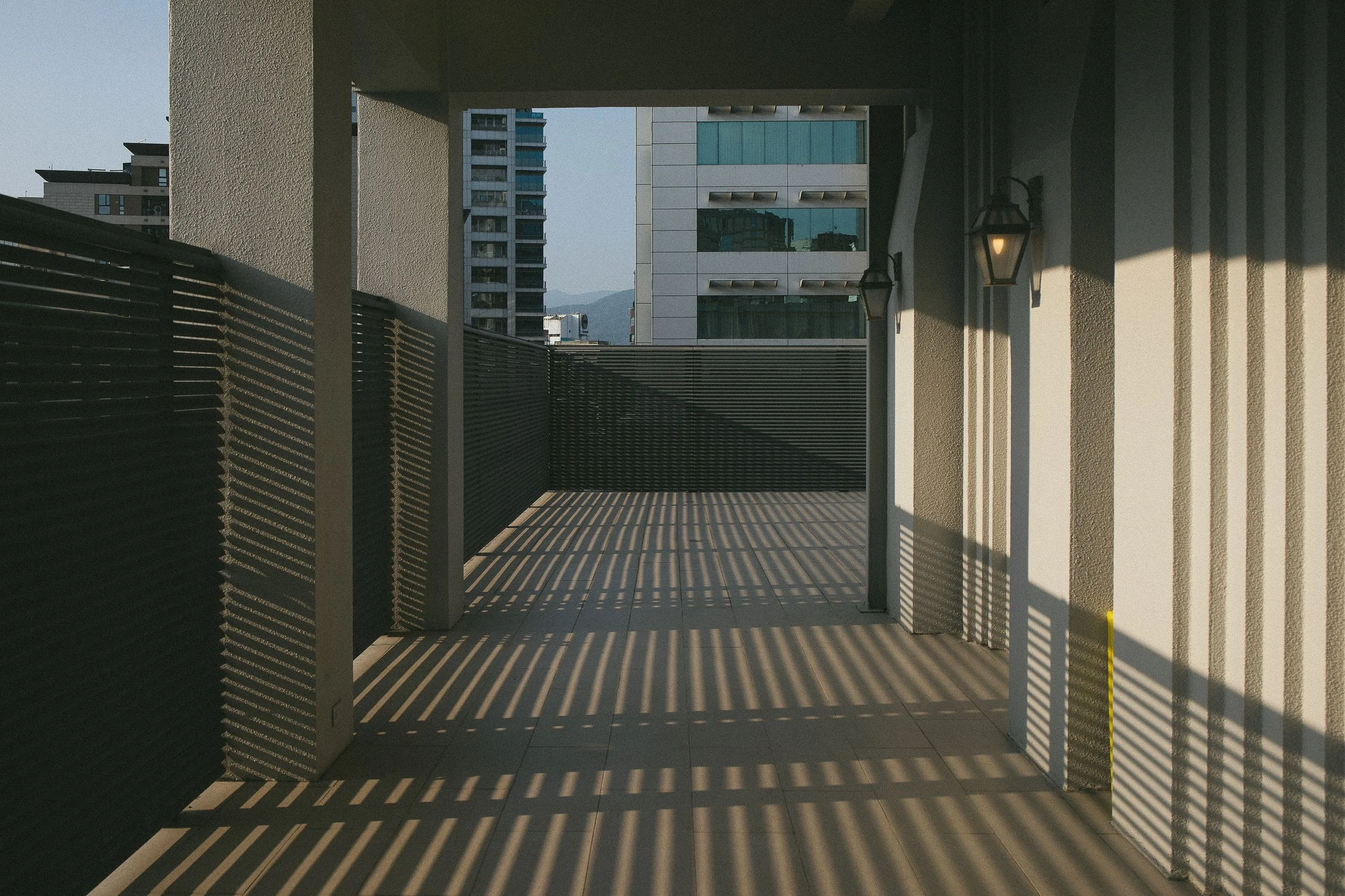 A balcony or terrace with shadows of horizontal slats cast on the floor, walls, and ceiling, overlooking modern high-rise buildings and a mountain in the background.