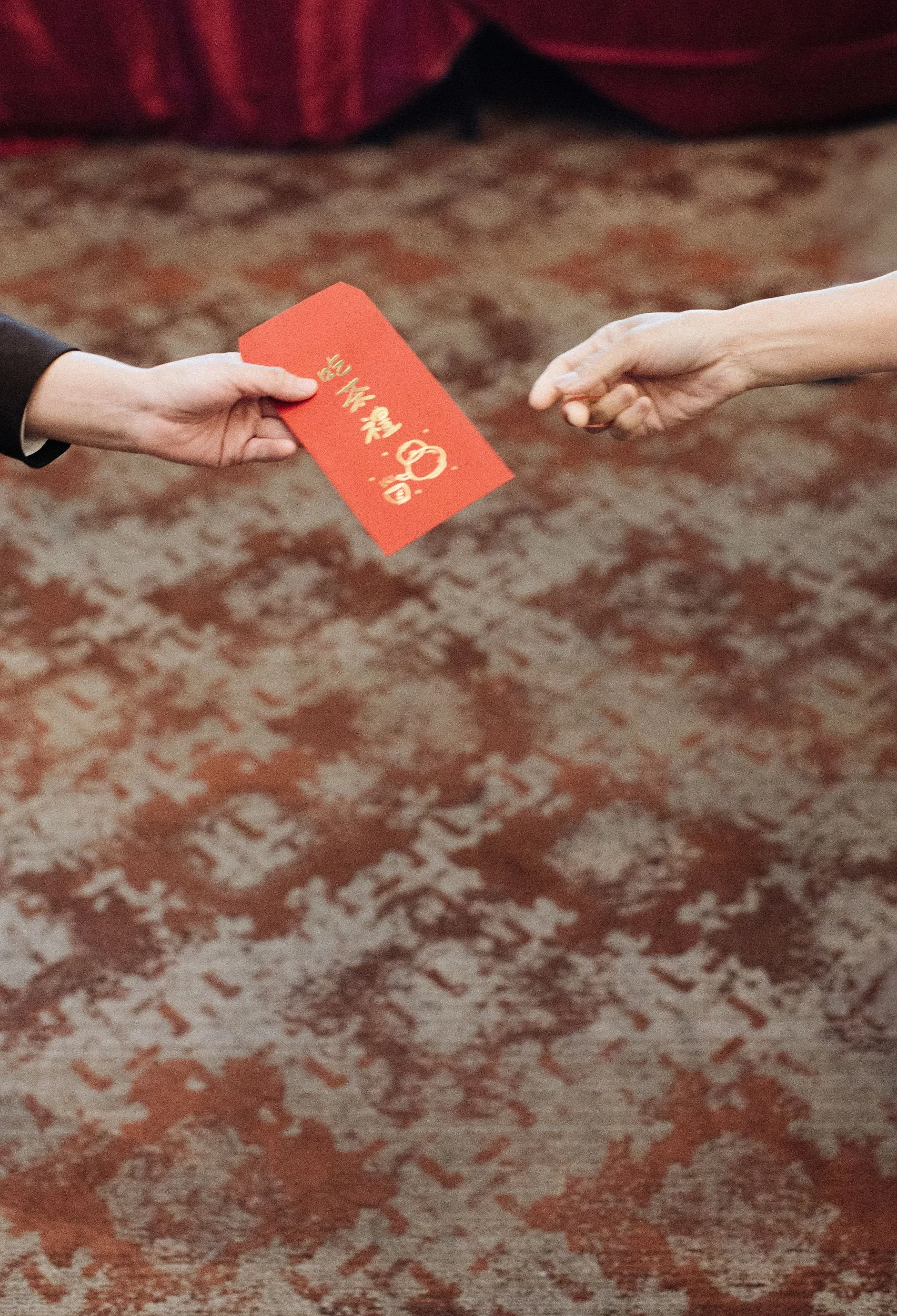A hand offering a red envelope to another hand, with a carpeted floor in the background.
