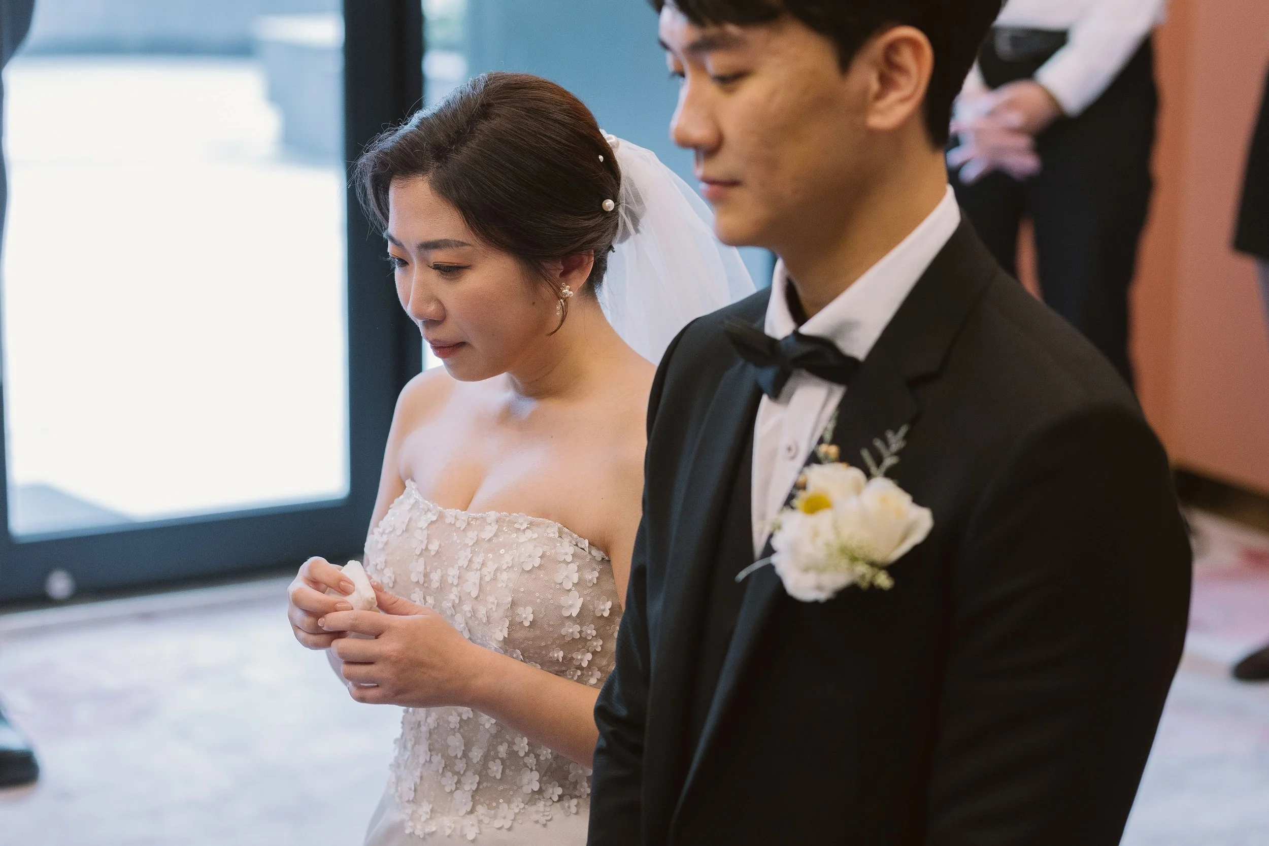 A bride and groom standing side by side during their wedding ceremony. The bride is wearing a white off-the-shoulder wedding gown with floral embellishments, and the groom is dressed in a black tuxedo with a white shirt and black bow tie. The bride h