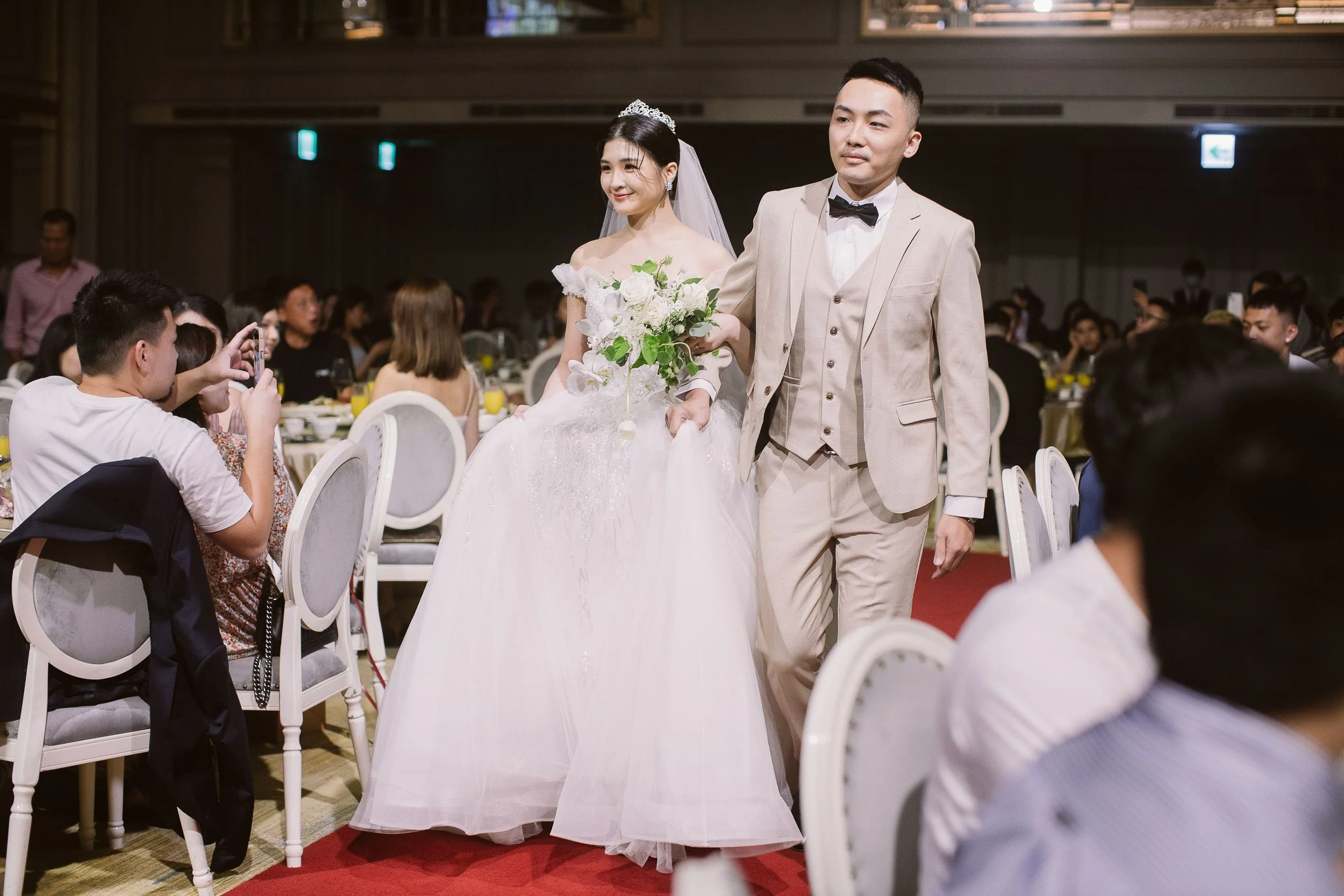 A bride and groom walking down the aisle at their wedding reception, with guests seated at tables watching and taking pictures.