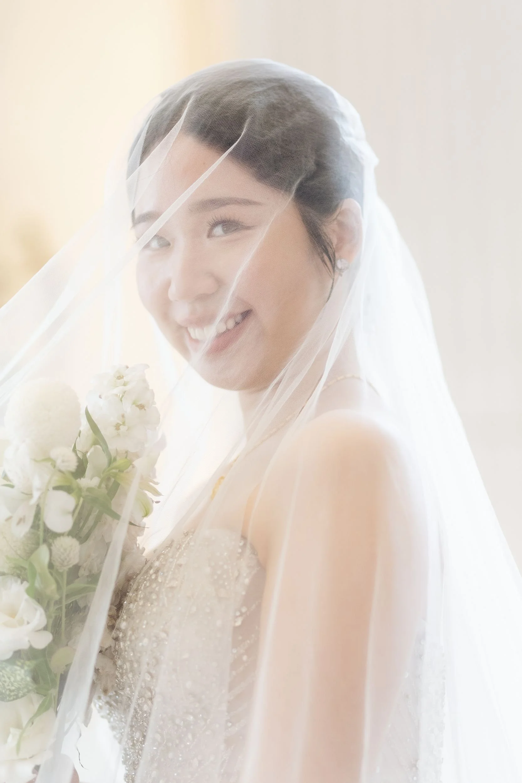 A woman smiling behind a sheer veil, holding a bouquet of white flowers, wearing a wedding dress with detailed embroidery.
