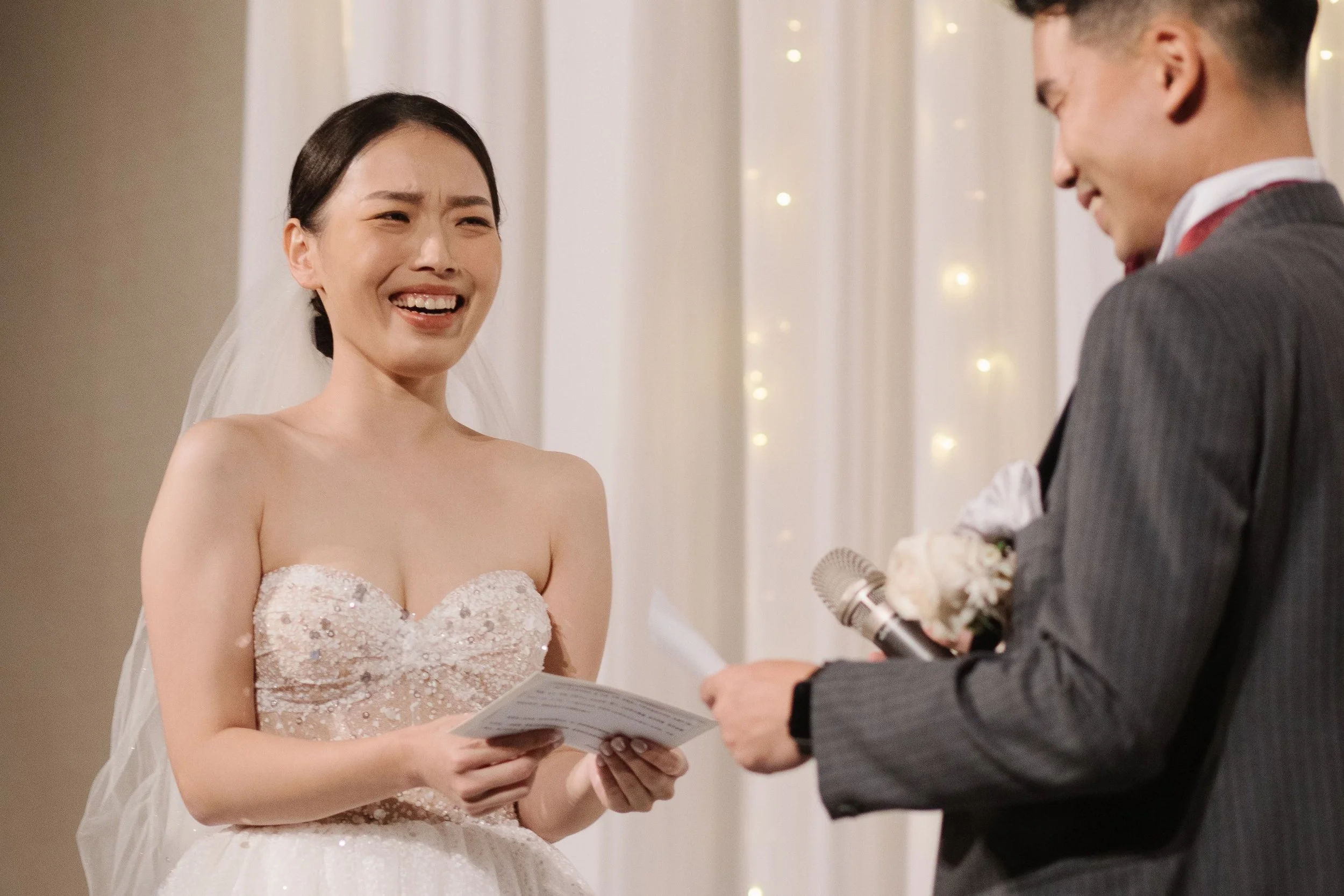 A bride and groom exchanging vows during a wedding ceremony indoors. The bride is smiling and holding a piece of paper, wearing a wedding dress with a veil, and the groom is dressed in a suit and tie, holding a microphone and possibly a ring or flowe