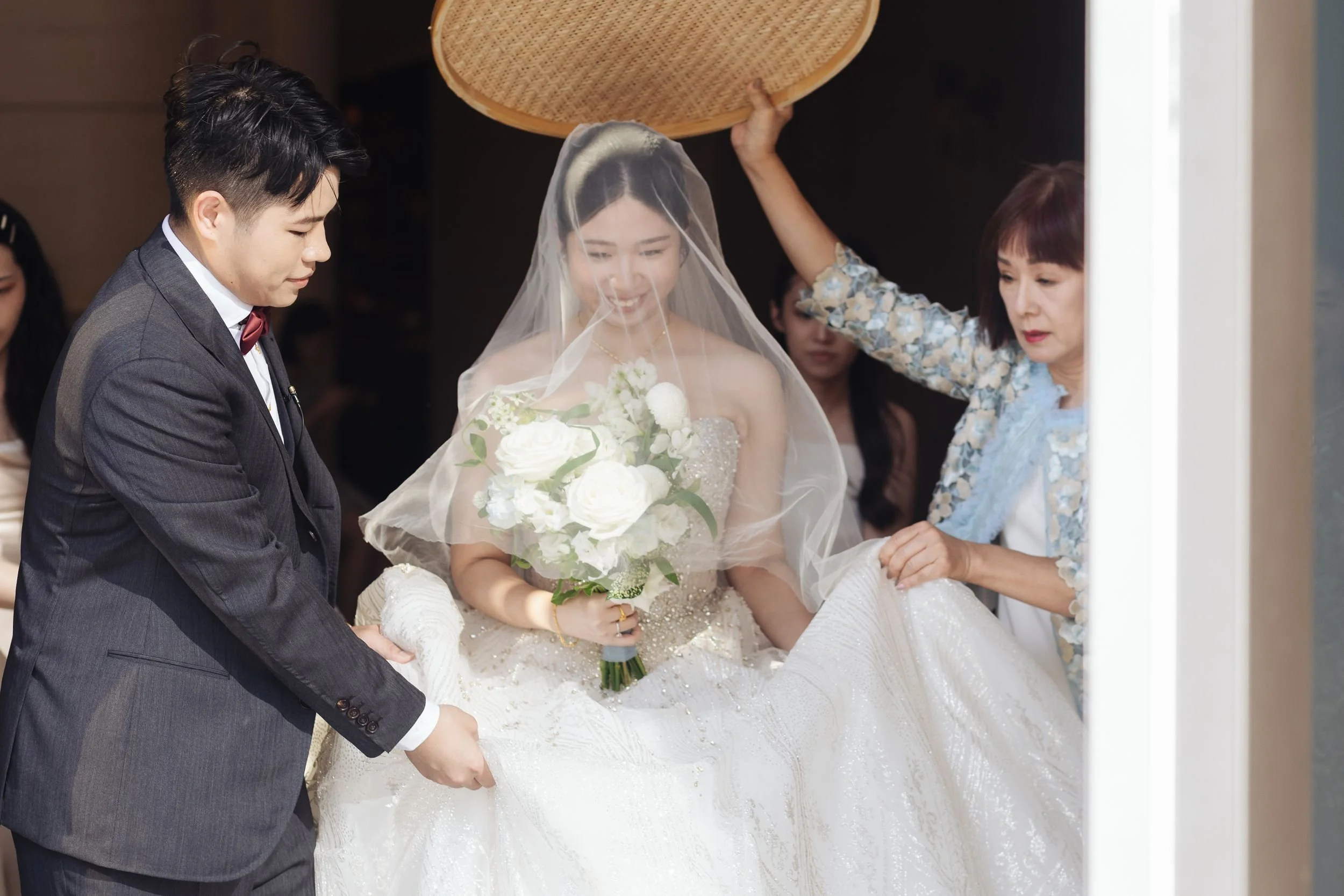 A bride in a white wedding dress and veil holding a bouquet of white flowers, surrounded by family members during a wedding ceremony.