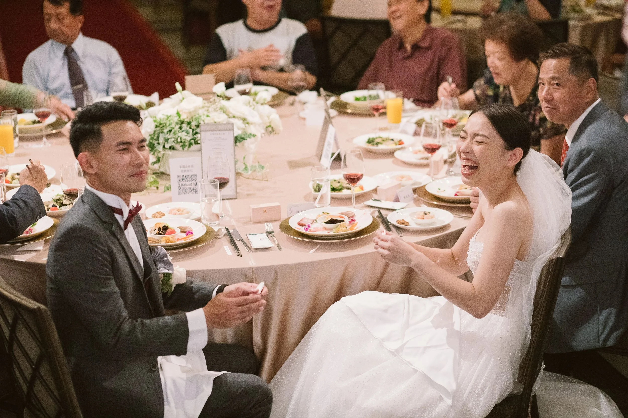 A wedding reception with a bride and groom sitting at a table surrounded by family members and friends. The bride is laughing and the groom is sitting beside her. The table is decorated with floral arrangements and set with plates of food, glasses, a