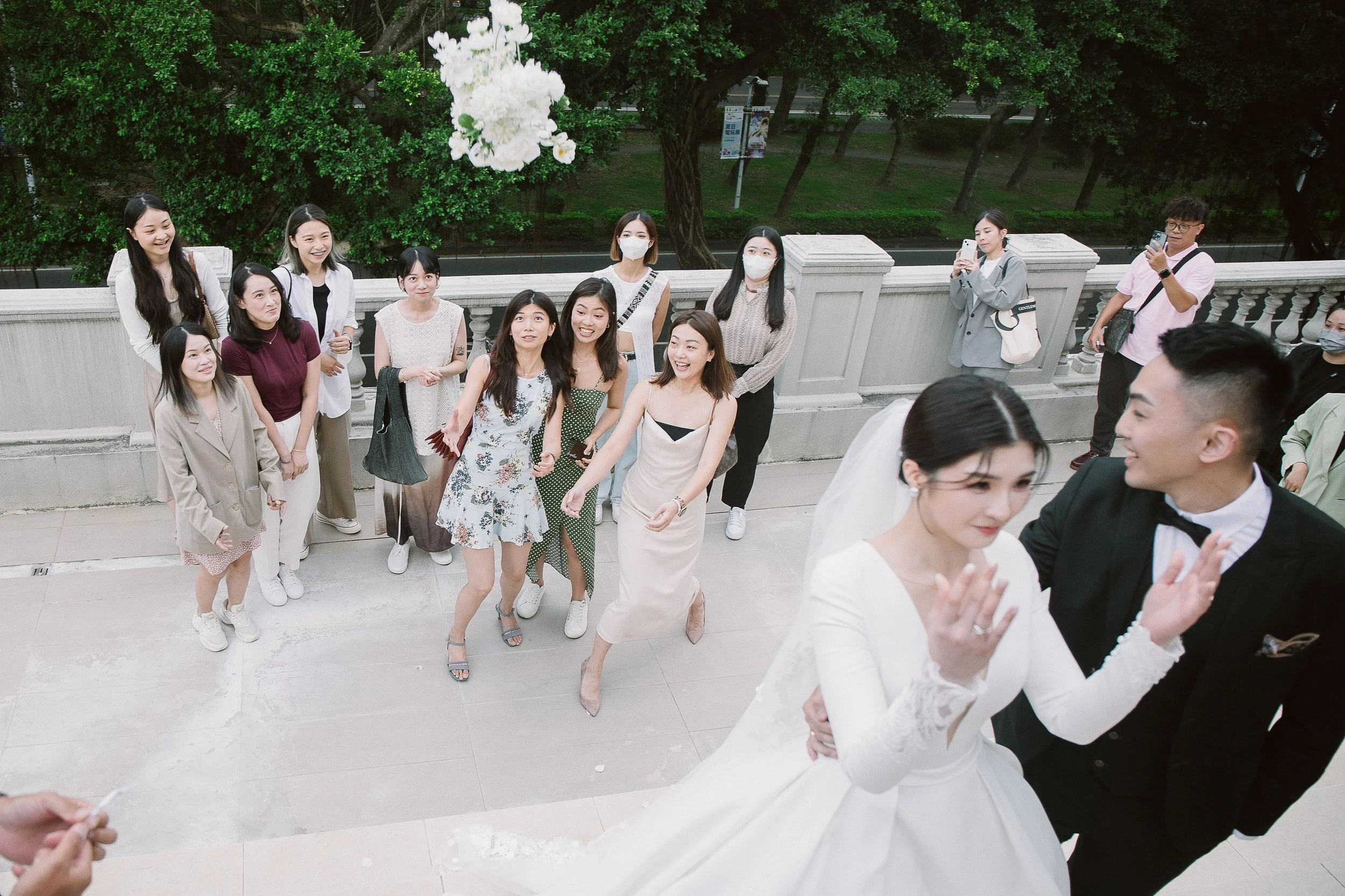 A group of women and a bride in wedding dress gathered outside on steps, celebrating wedding, some wearing masks, others taking photos, with green trees in background.
