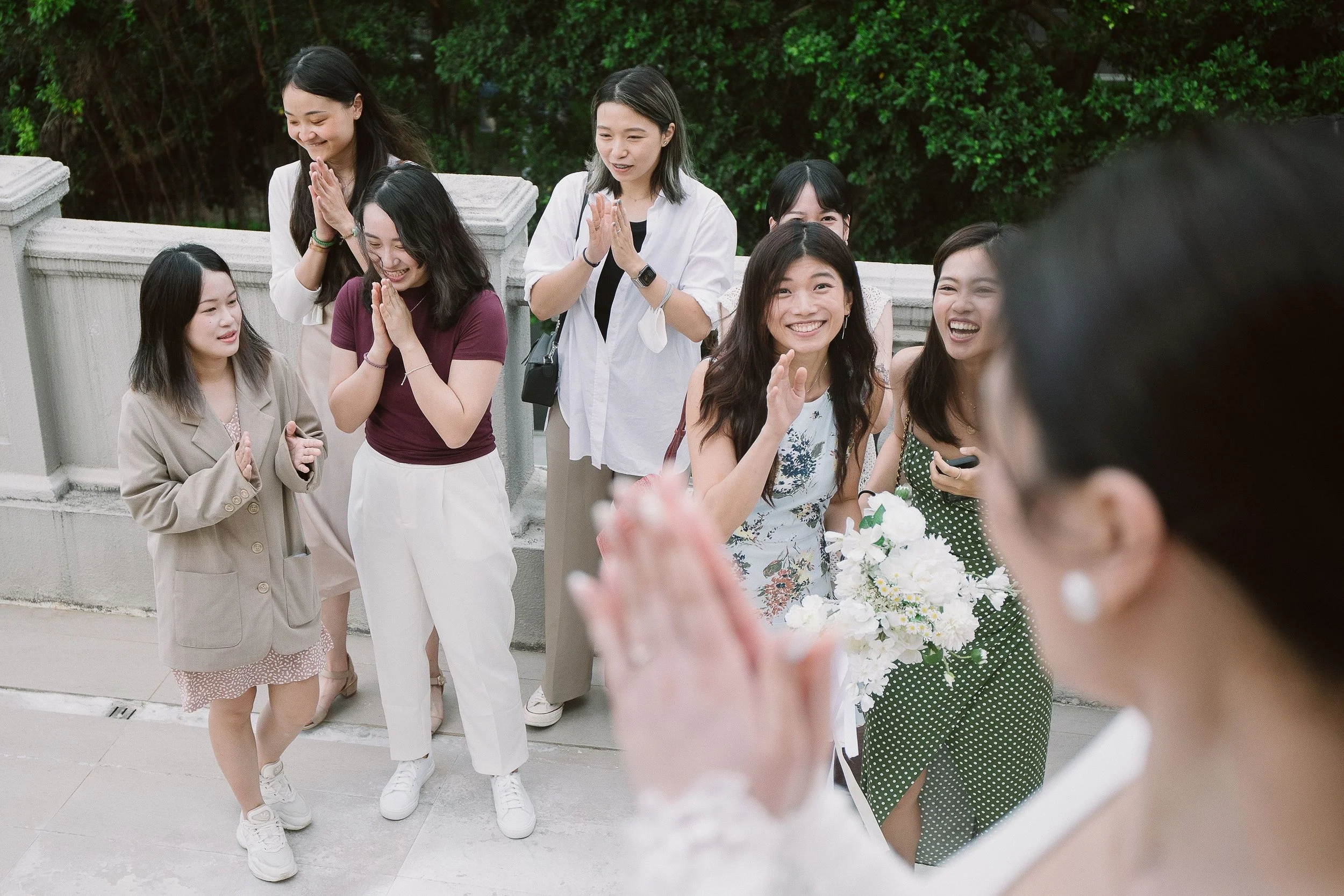 Group of women smiling and greeting each other outdoors, some with hands clasped in greeting and others holding flowers, in front of a green hedgerow.