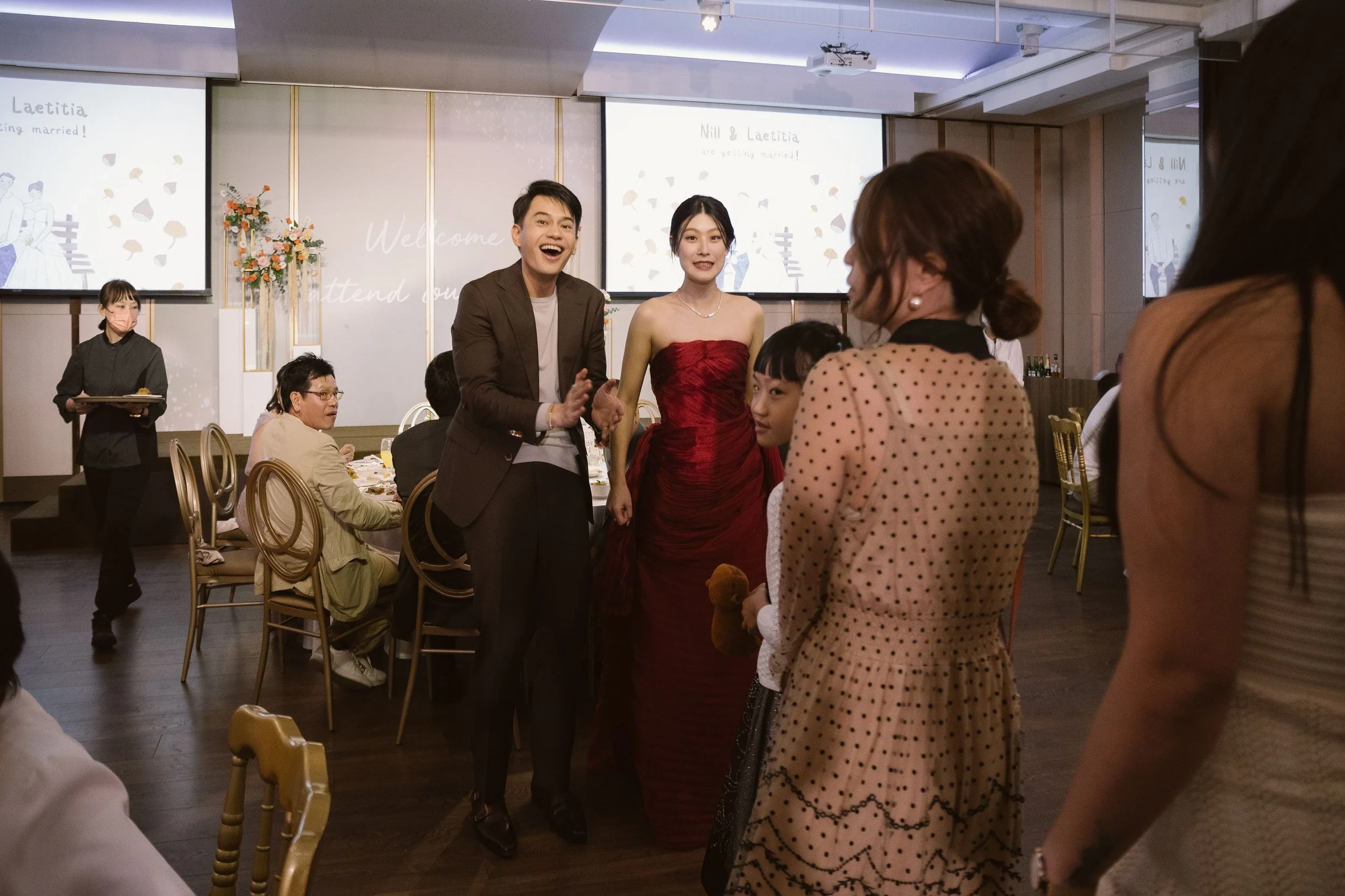 A wedding reception with a bride and groom holding hands, smiling, and surrounded by guests. The bride is wearing a red dress and the groom is in a brown suit. There are large screens in the background displaying wedding graphics and text.